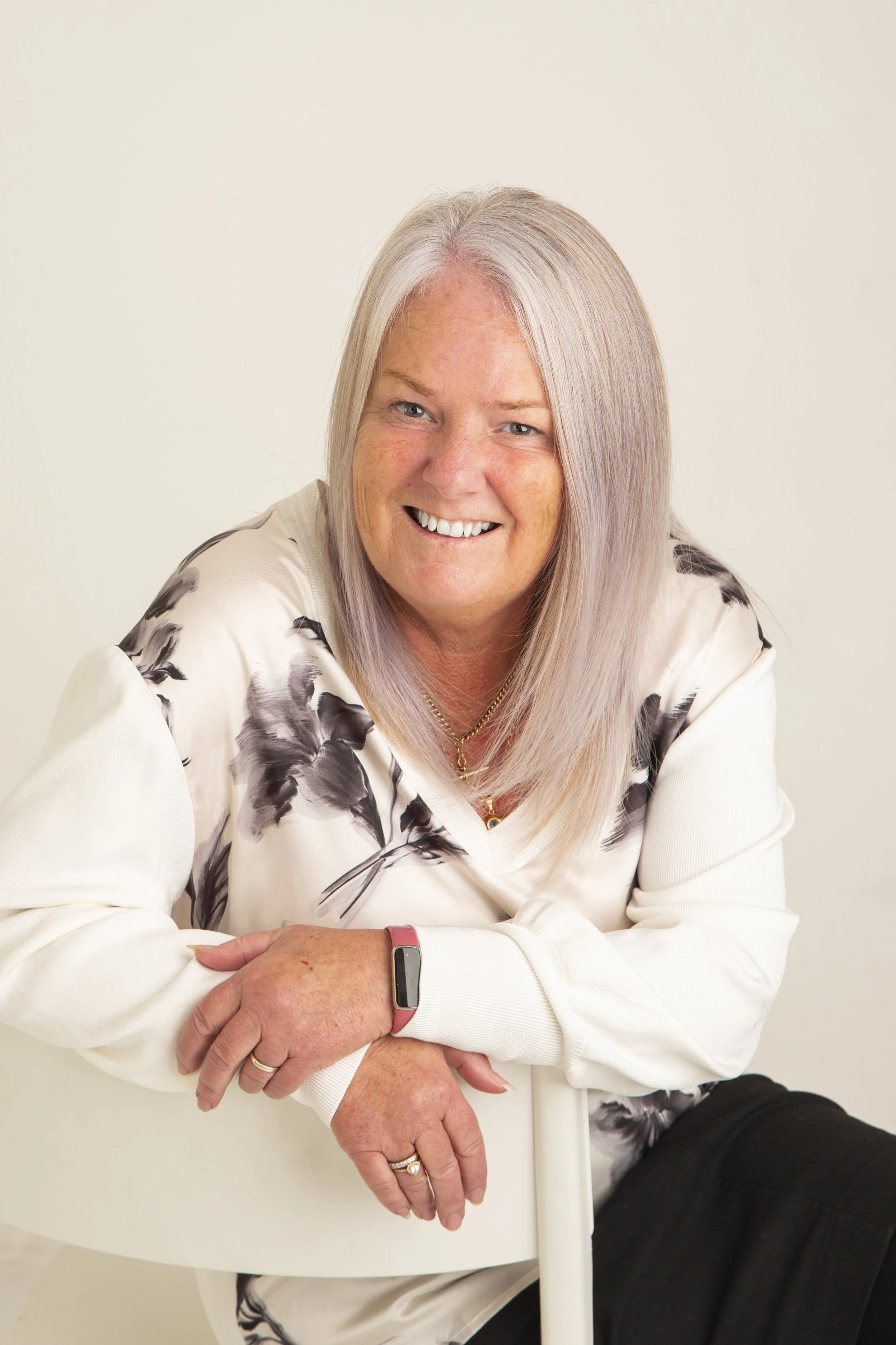 A smiling woman with long blonde hair, wearing a white top with black floral pattern, sitting on a white chair against a plain beige background.