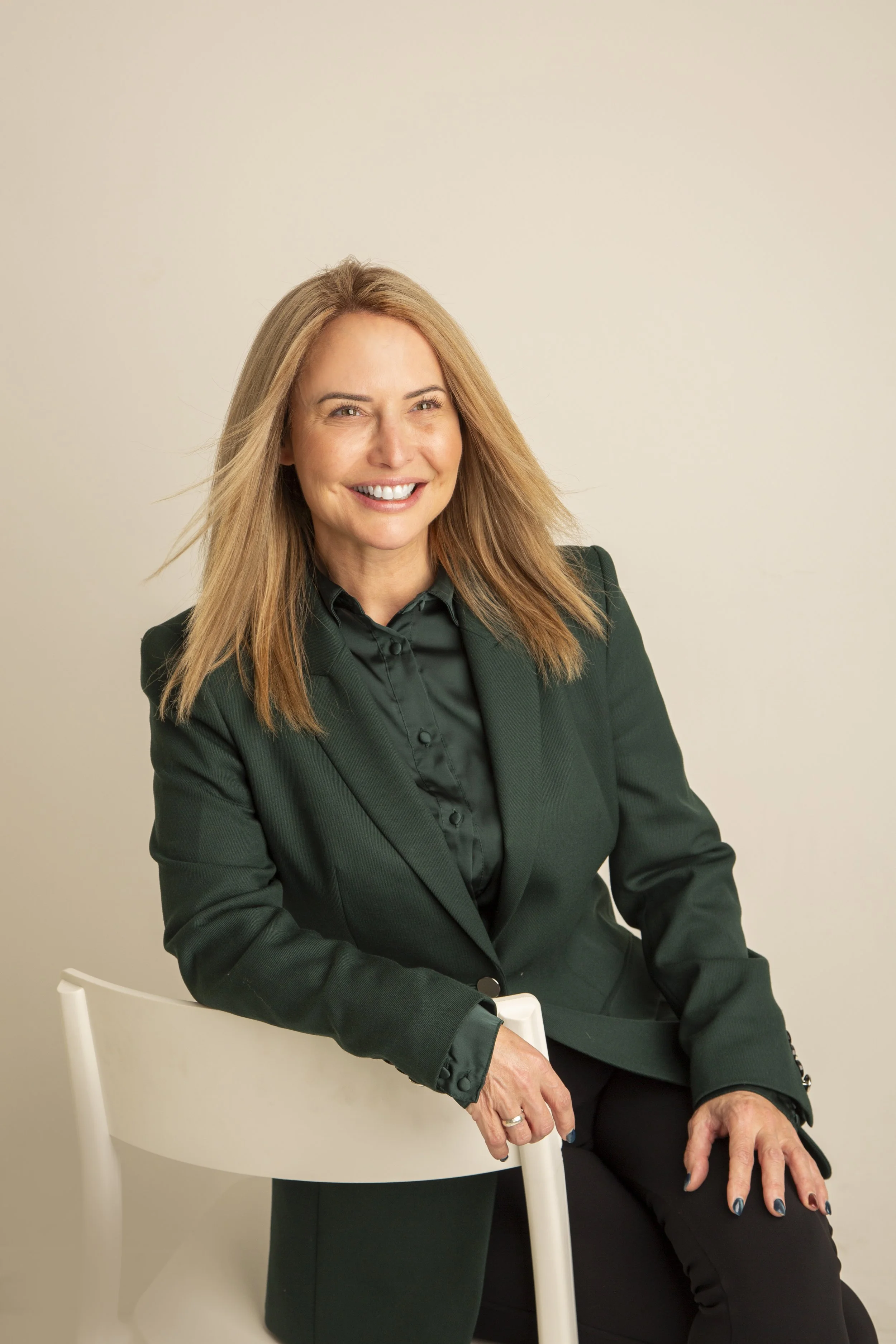 A smiling woman with shoulder-length blond hair, wearing a dark green blazer and black shirt, sitting on a white chair against a plain beige background.