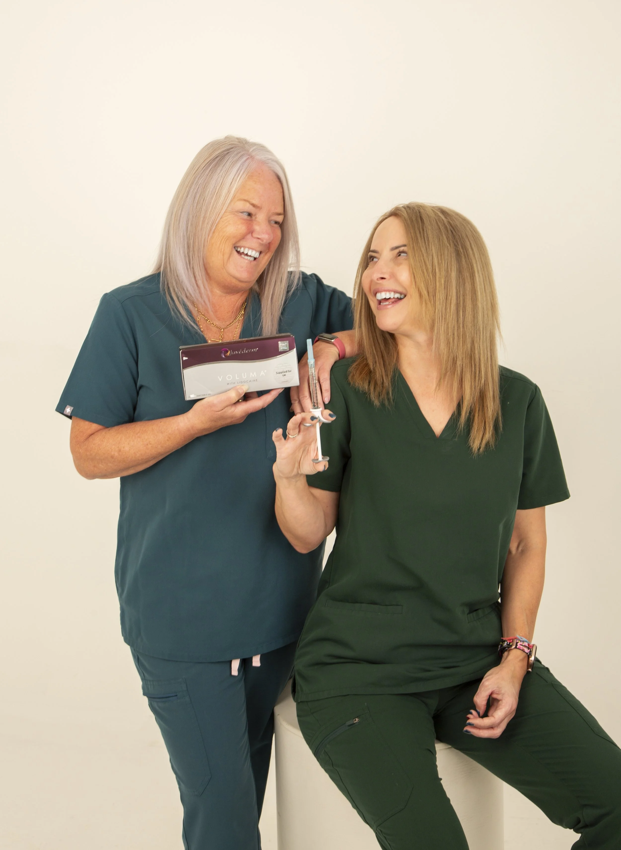 Two women in medical scrubs smiling and laughing, one holding a syringe and the other holding a box of Juvederm cosmetic filler.