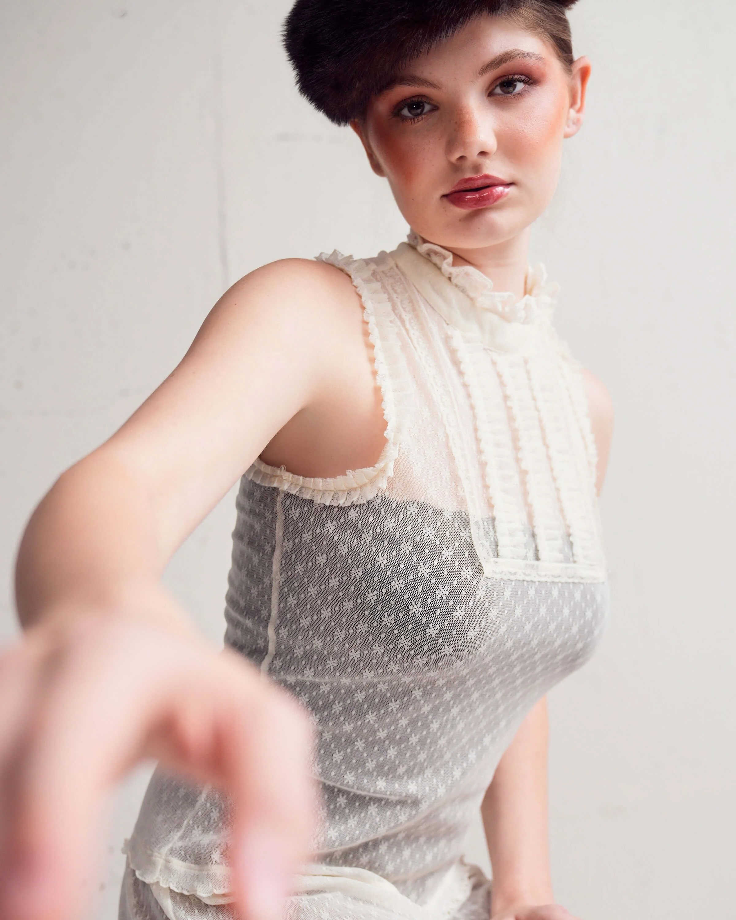 Close-up of a young woman with short dark hair, wearing a sleeveless cream-colored top with ruffled details, looking at the camera with a neutral expression, standing against a plain light background.