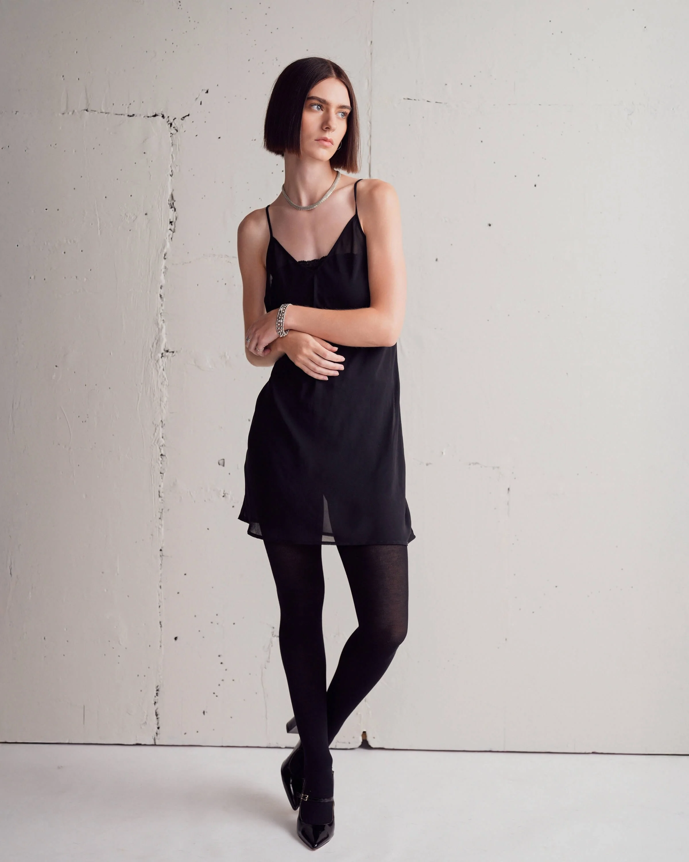 A young woman with shoulder-length dark hair, wearing a black spaghetti strap dress, black tights, and black heels, stands with crossed arms against a plain, light-colored textured wall.