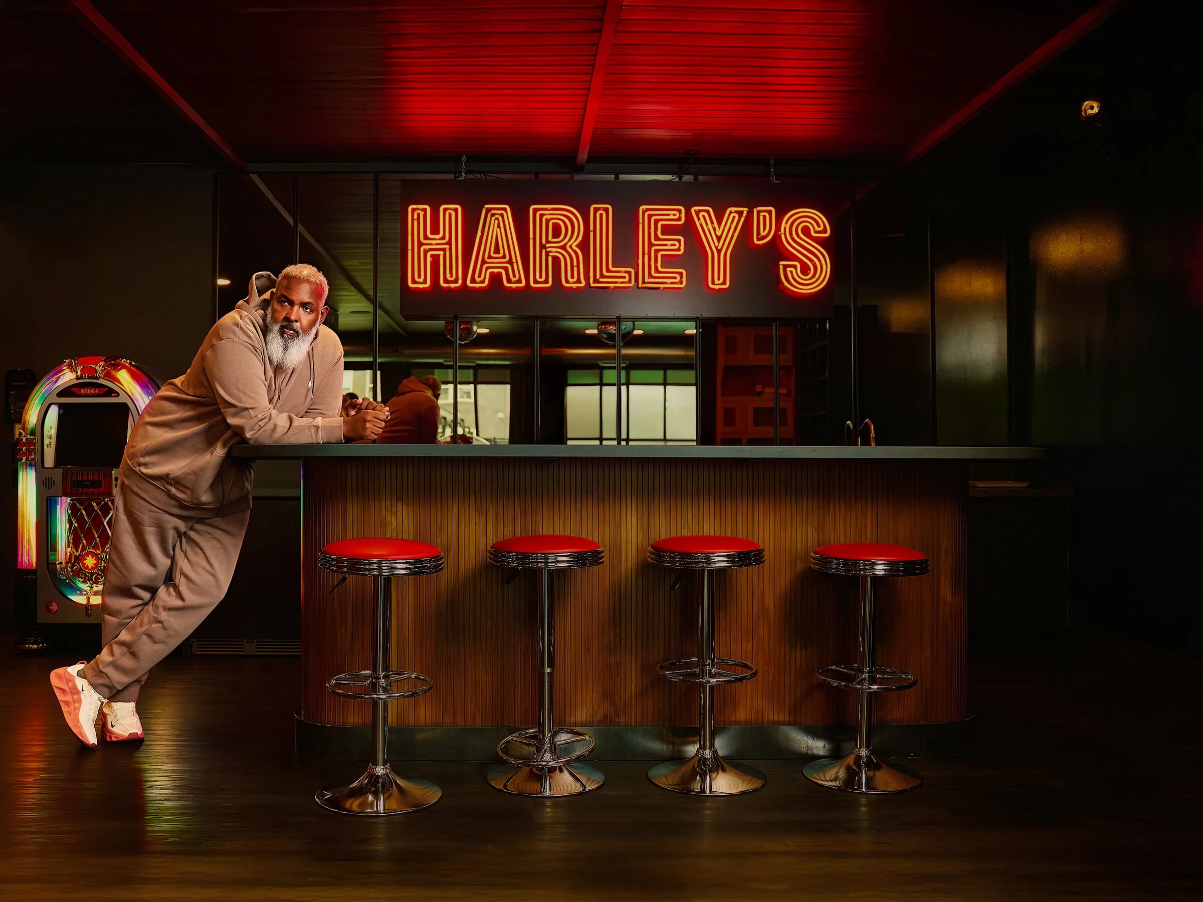 A man leaning on a bar counter with a neon 'Harley's' sign in the background and four red-cushioned bar stools in front of him, inside a dimly lit bar or club.