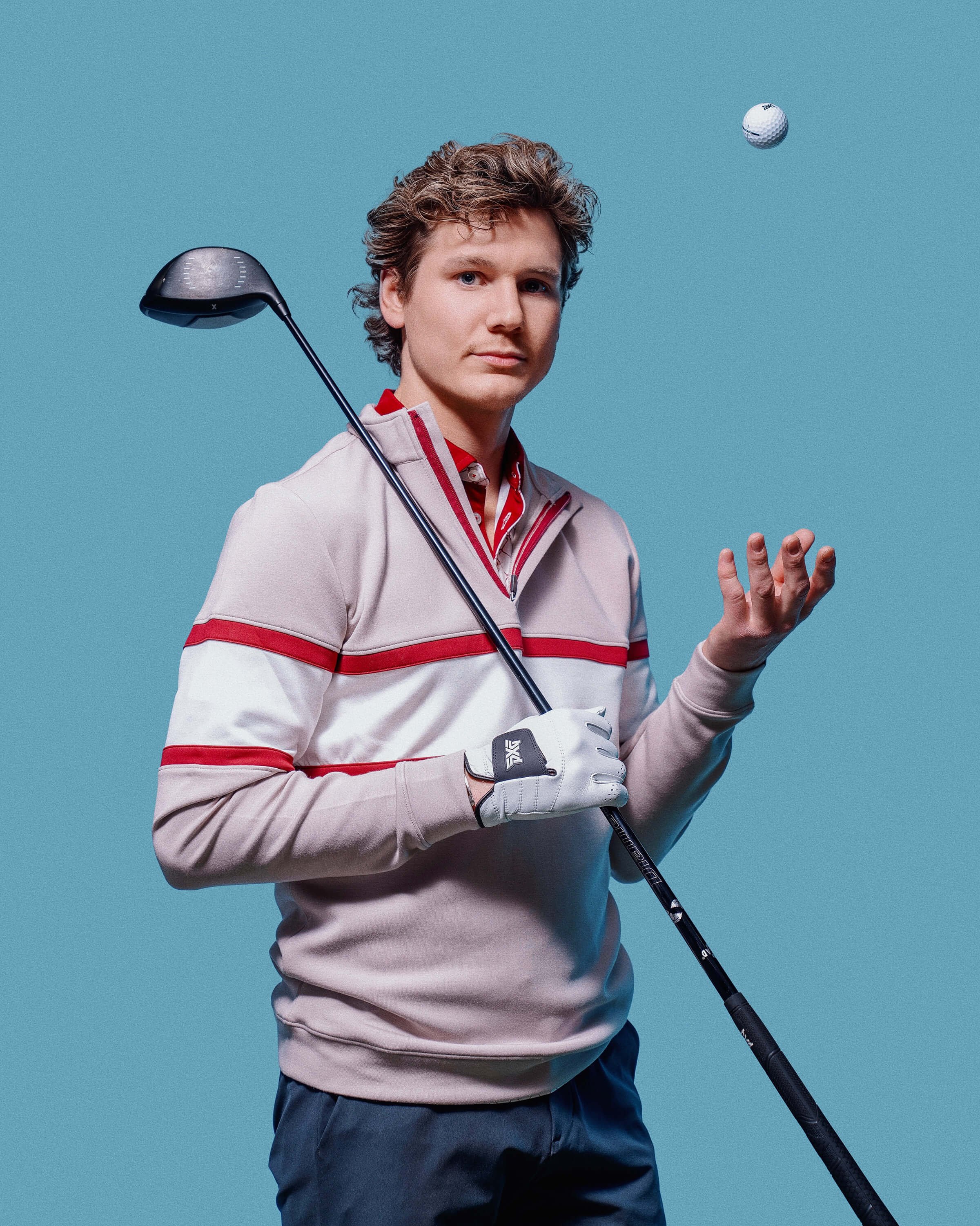 A young man with wavy brown hair stands against a light blue background, holding a golf club in his right hand and balancing a golf ball in the air with his left hand.