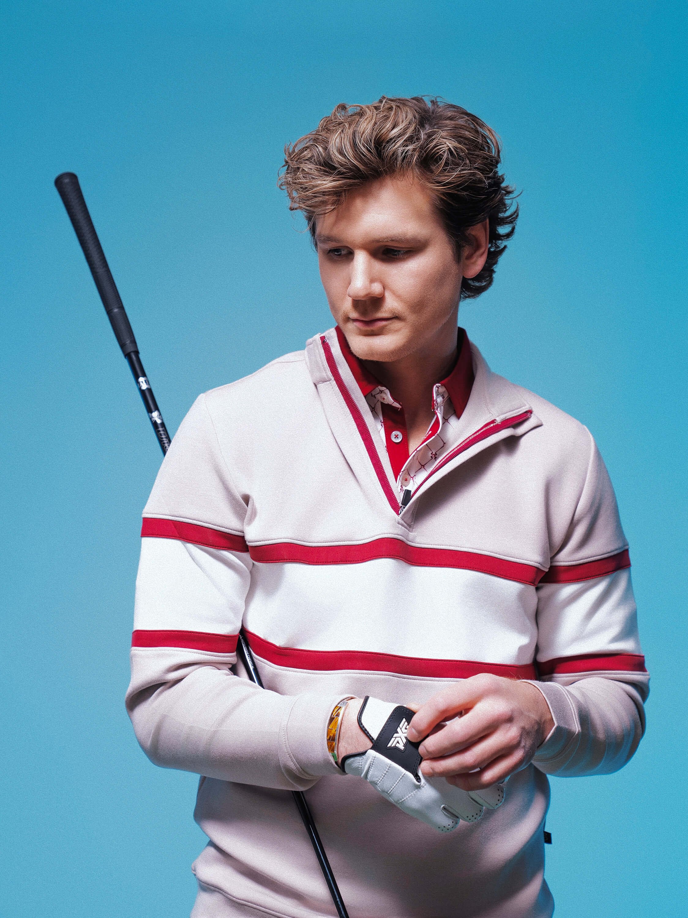 A young man in a beige and red striped golf sweater and a white glove, holding a golf club, standing against a blue background.