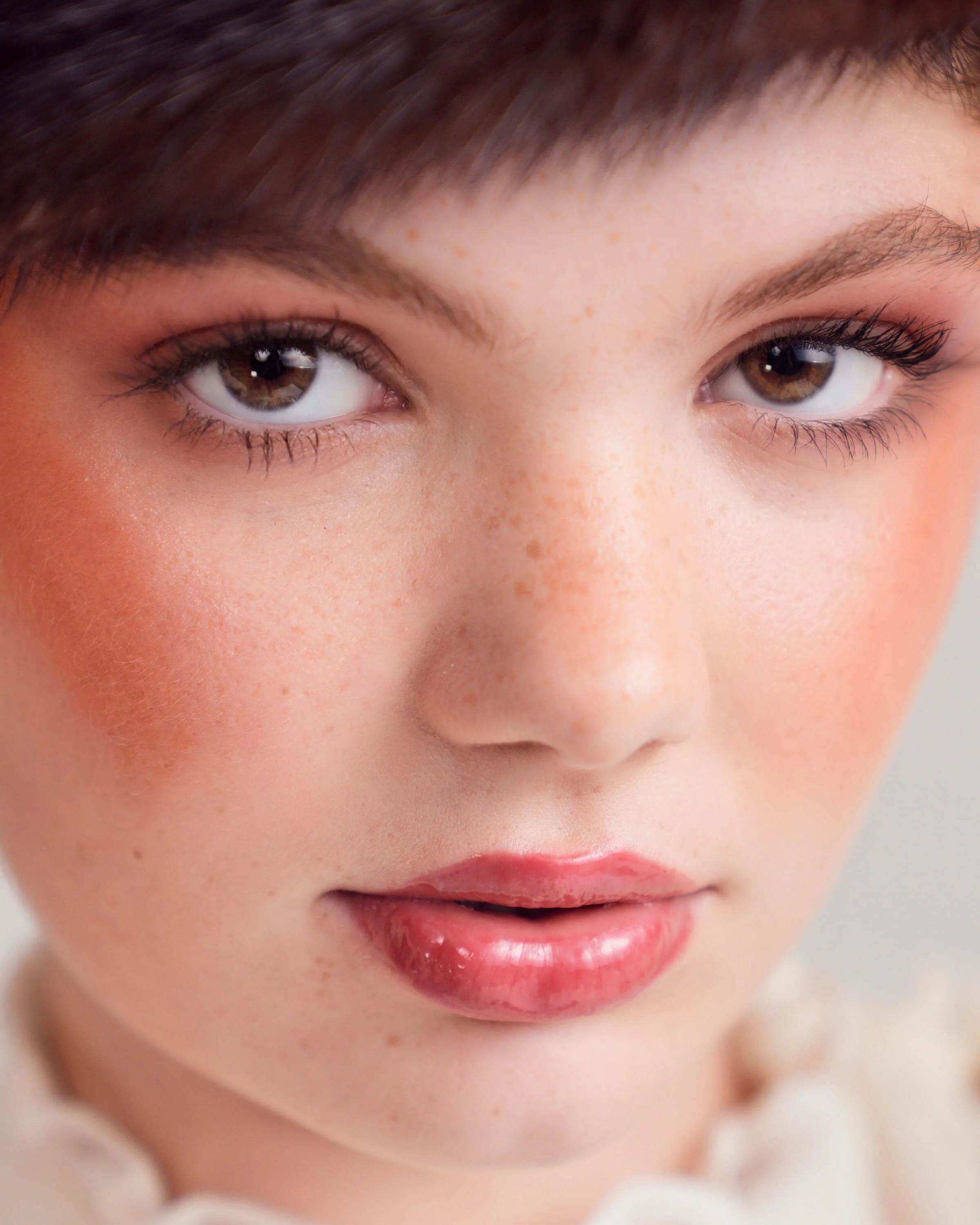 Close-up of a young woman's face with brown eyes, freckles, pink makeup, and natural lips.