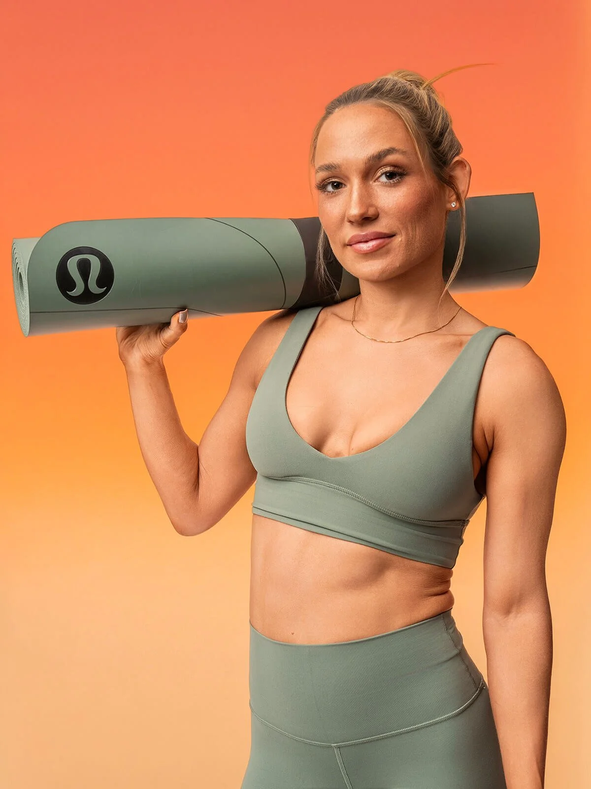 A woman in workout attire holding a rolled yoga mat on her shoulder against a warm orange background.