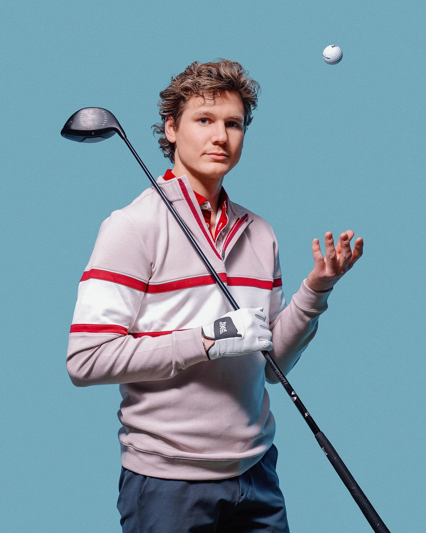 A young man in a gray and red golf sweater holding a golf club, with a golf ball in the air against a blue background.