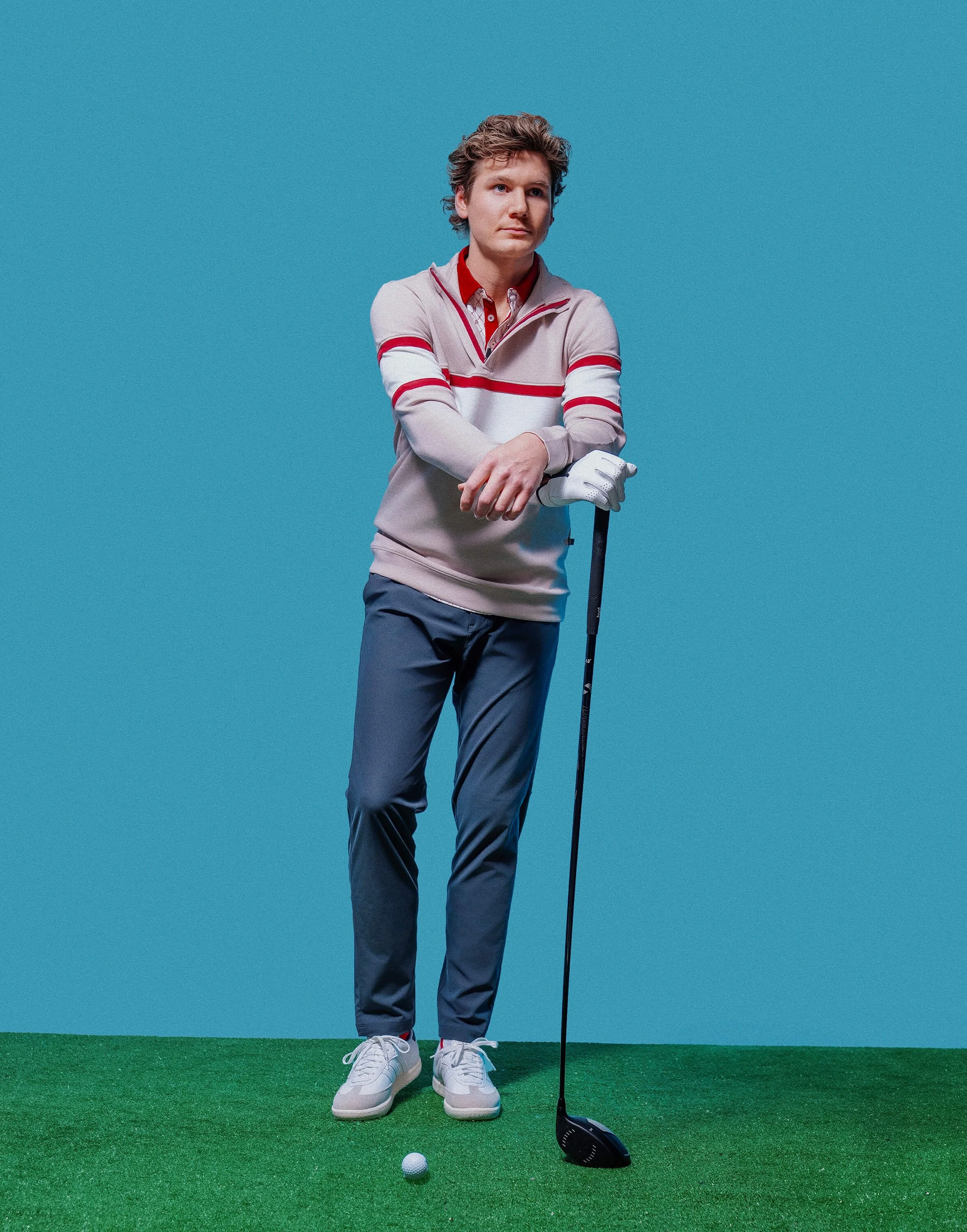 A young man in a beige, red, and white sweater stands on a green artificial turf, holding a golf club, with a white golf ball in front of his shoes, against a plain blue background.