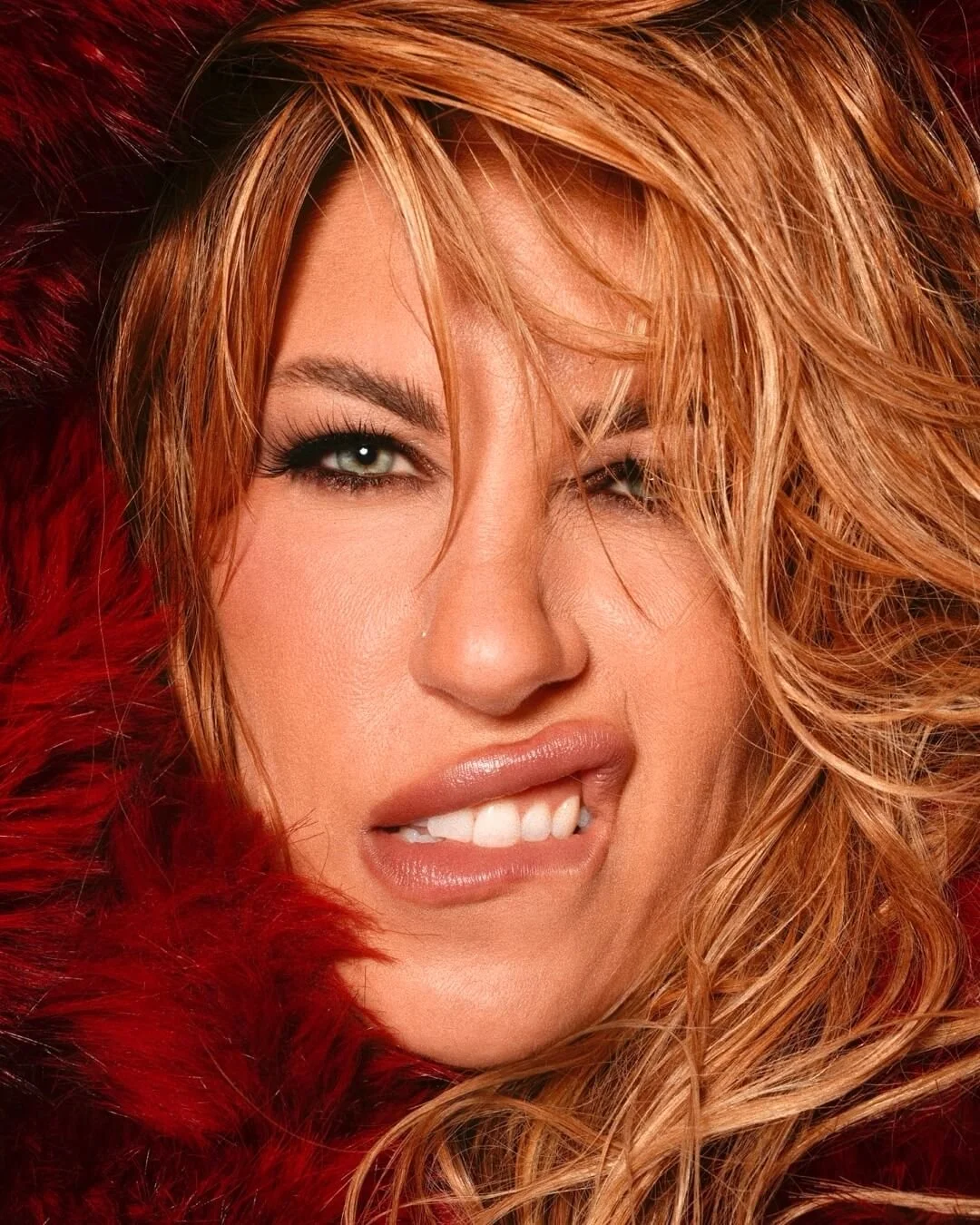 Close-up of a woman with long, wavy, reddish-blonde hair, one eye winking, and wearing red fur around her shoulders.
