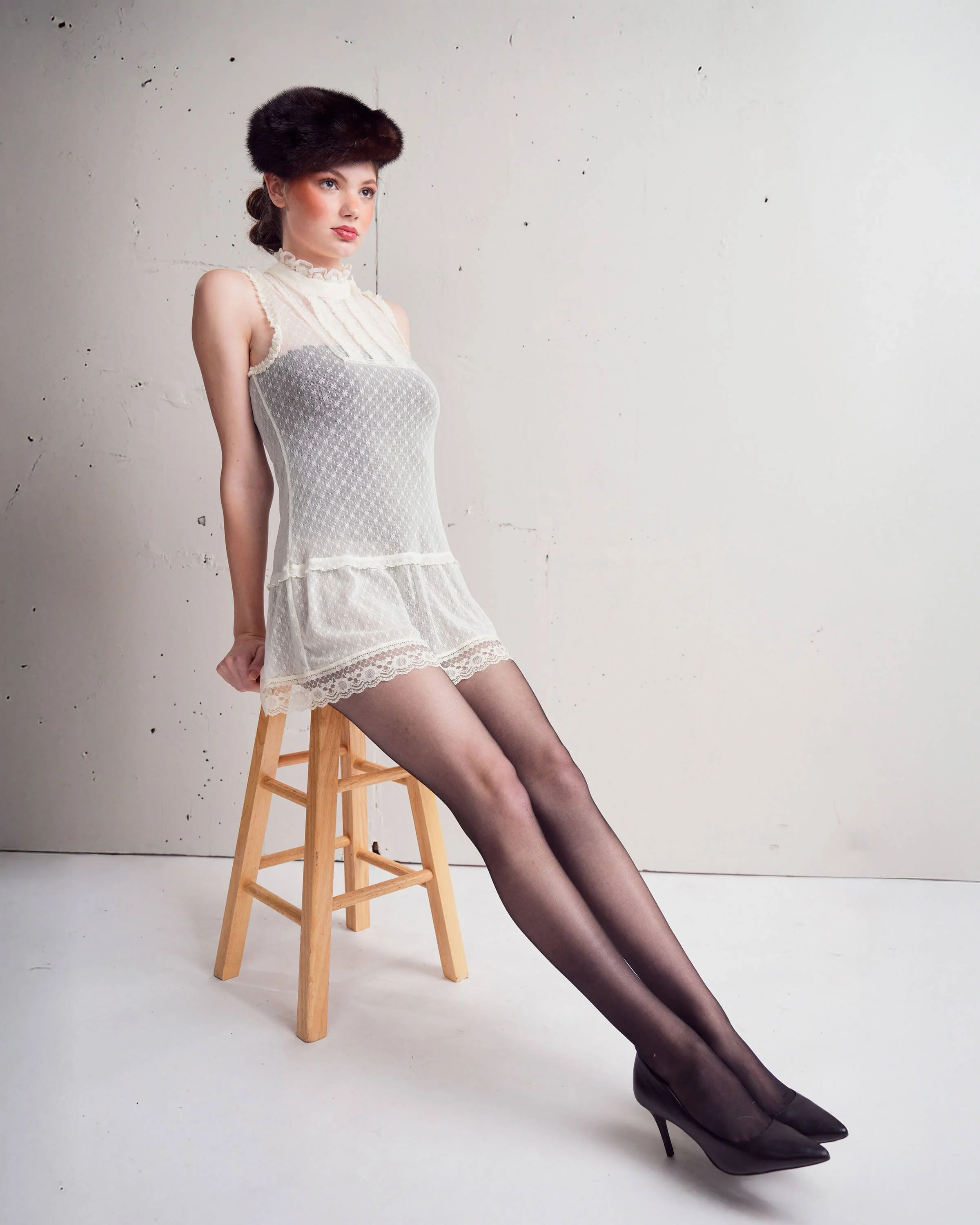 Woman wearing sheer white dress, black sheer stockings, black heels, and a dark fur hat sitting on a wooden stool against a plain wall.