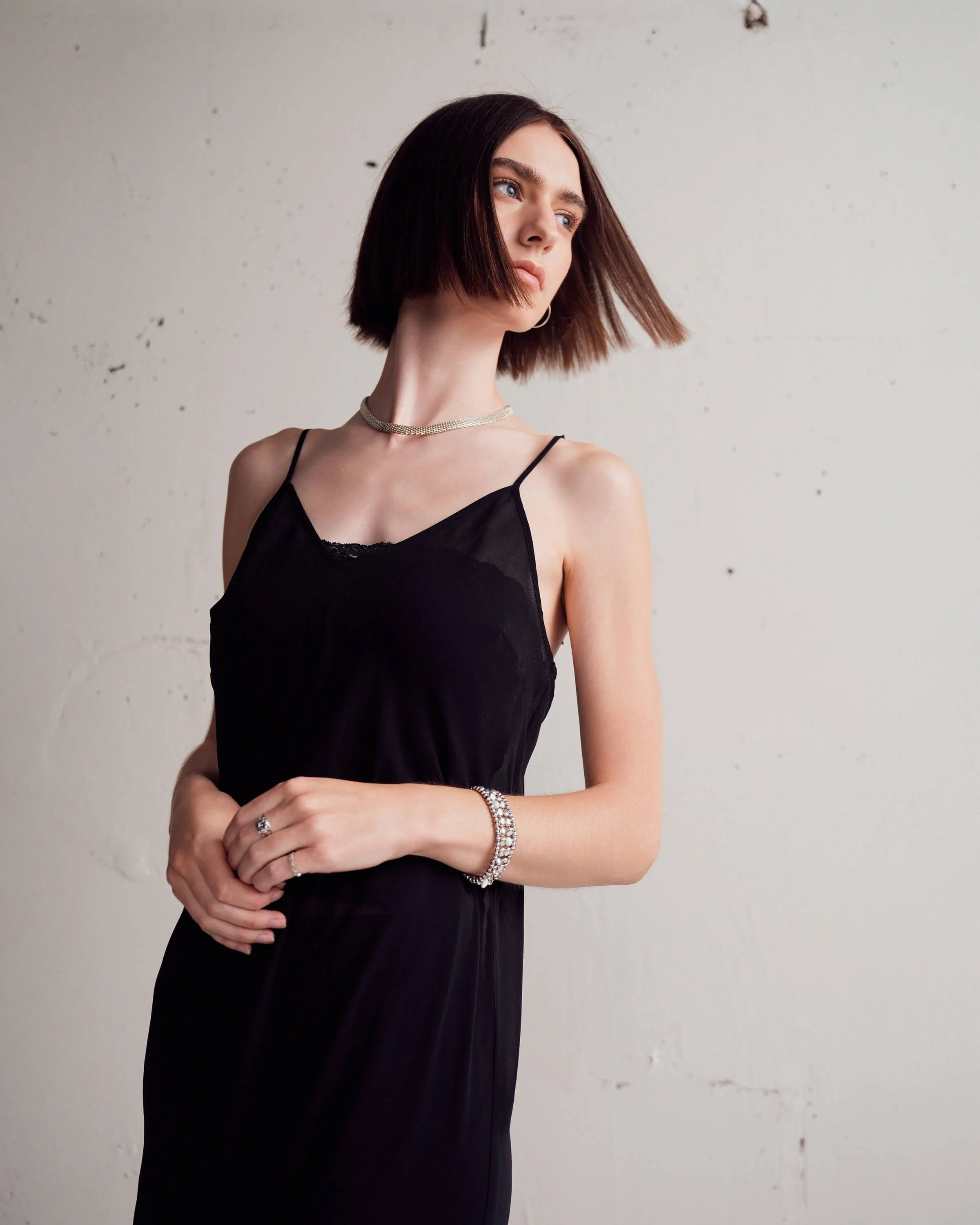 A woman with shoulder-length brown hair wearing a black spaghetti strap dress, a silver choker necklace, and a bracelet, standing against a plain white wall.