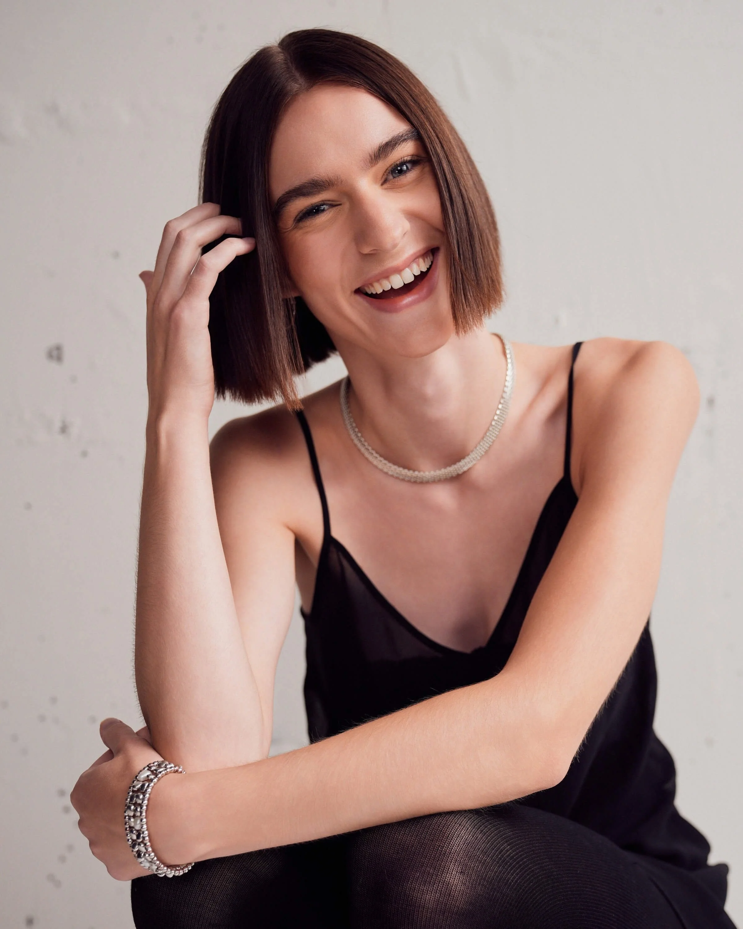 A smiling young woman with short brown hair wearing a black dress, pearl necklace, and silver jewelry, sitting against a plain white wall.