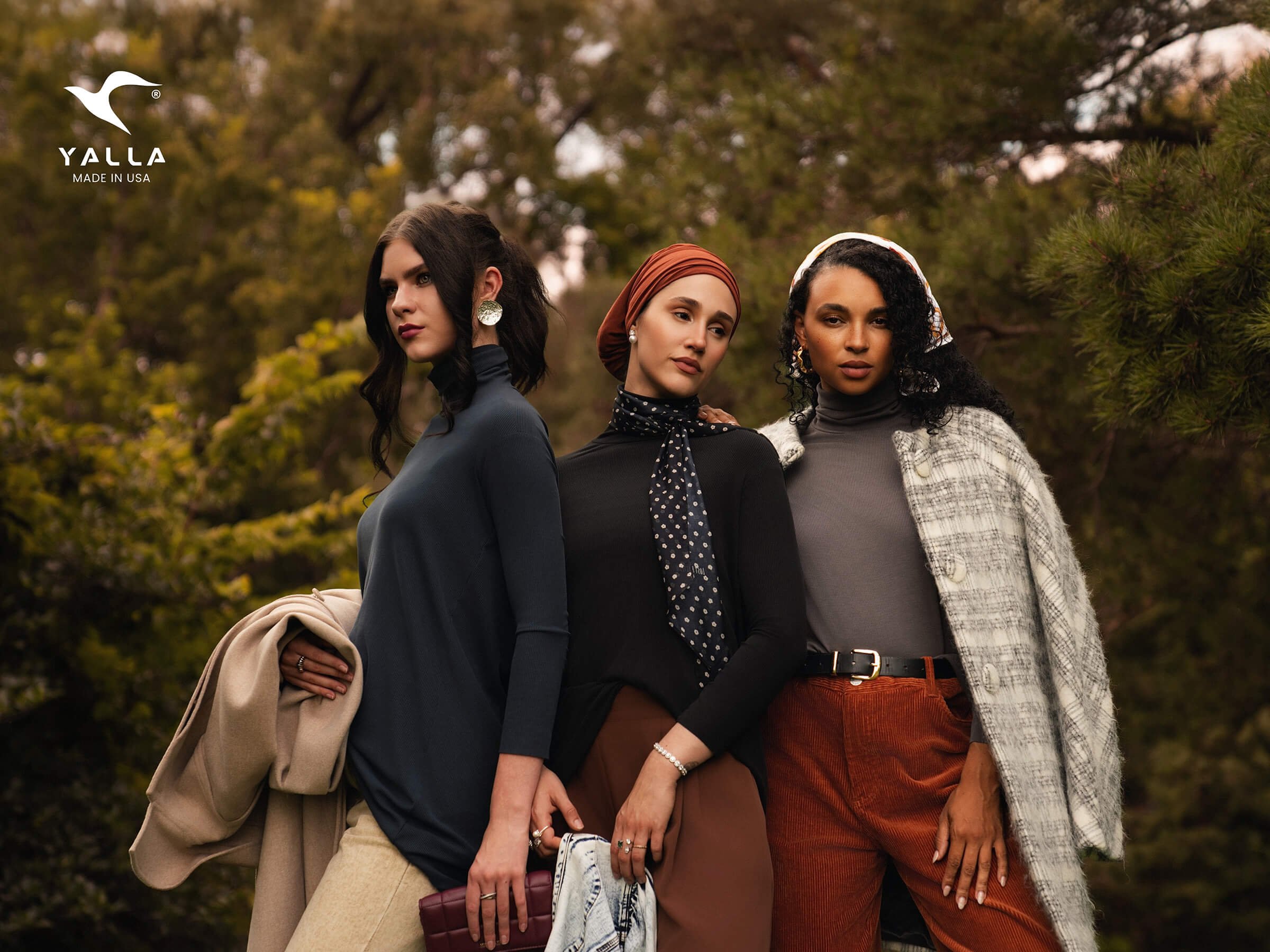 Three women standing outdoors among trees, dressed in stylish autumn clothing, posing confidently.
