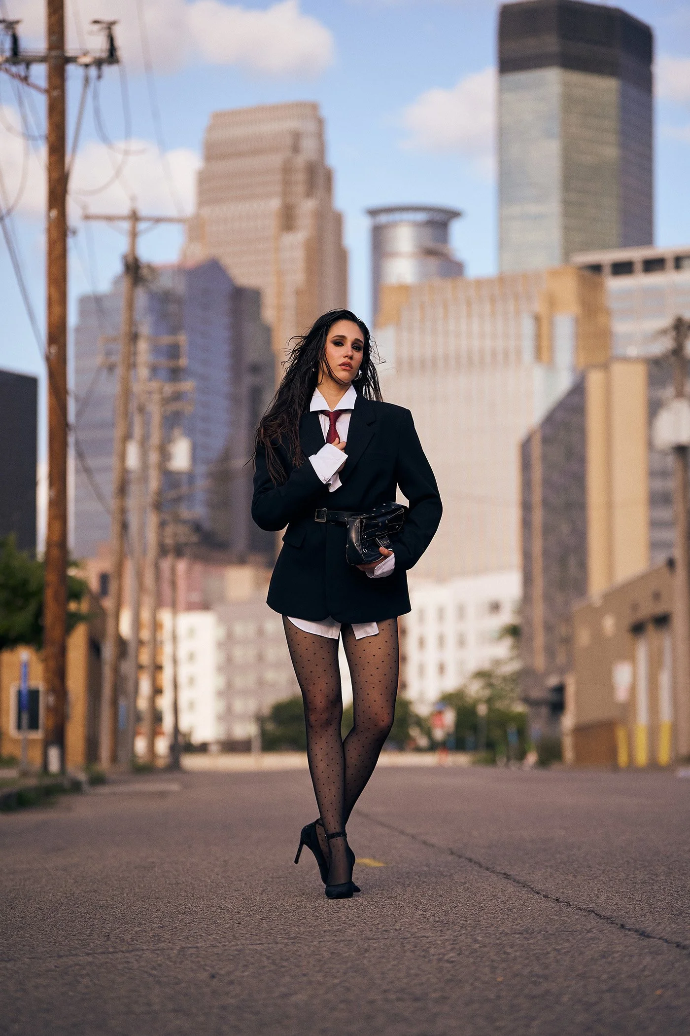 Editorial fashion photograph of a woman in a blazer and heels standing on a city street with modern buildings, styled for a commercial fashion campaign in downtown Minneapolis