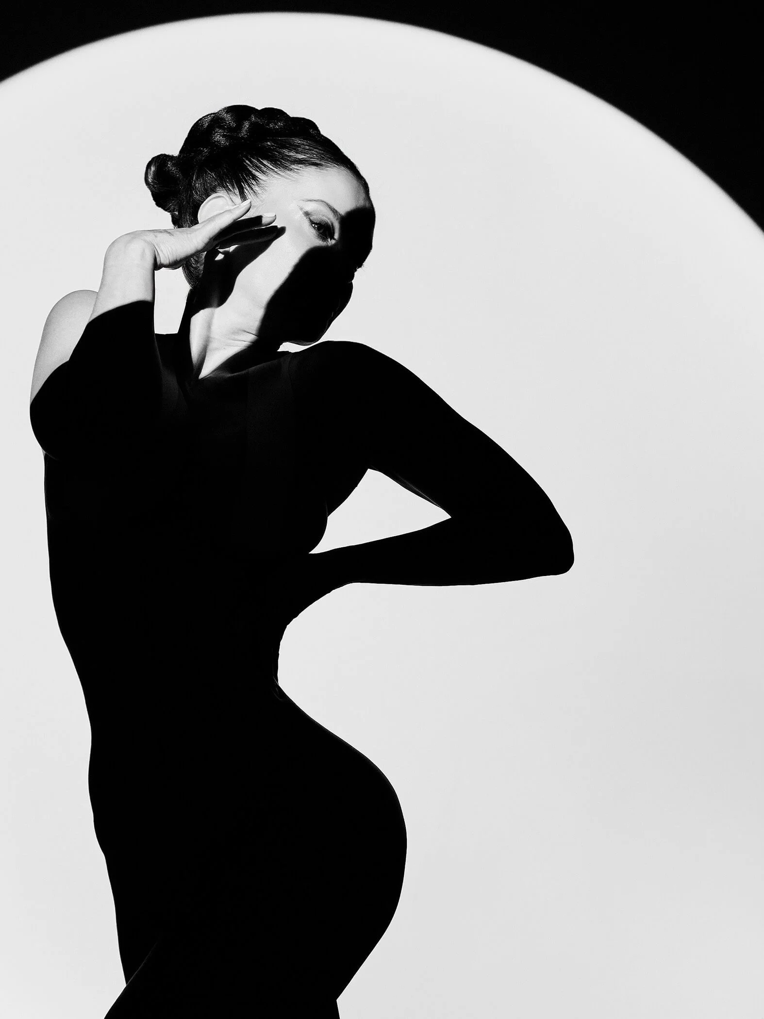 Black and white photograph of a woman in a form-fitting dress, posing with her hand near her face and her body turned slightly to the side, with a stark contrasting background and a semi-circular white backdrop.