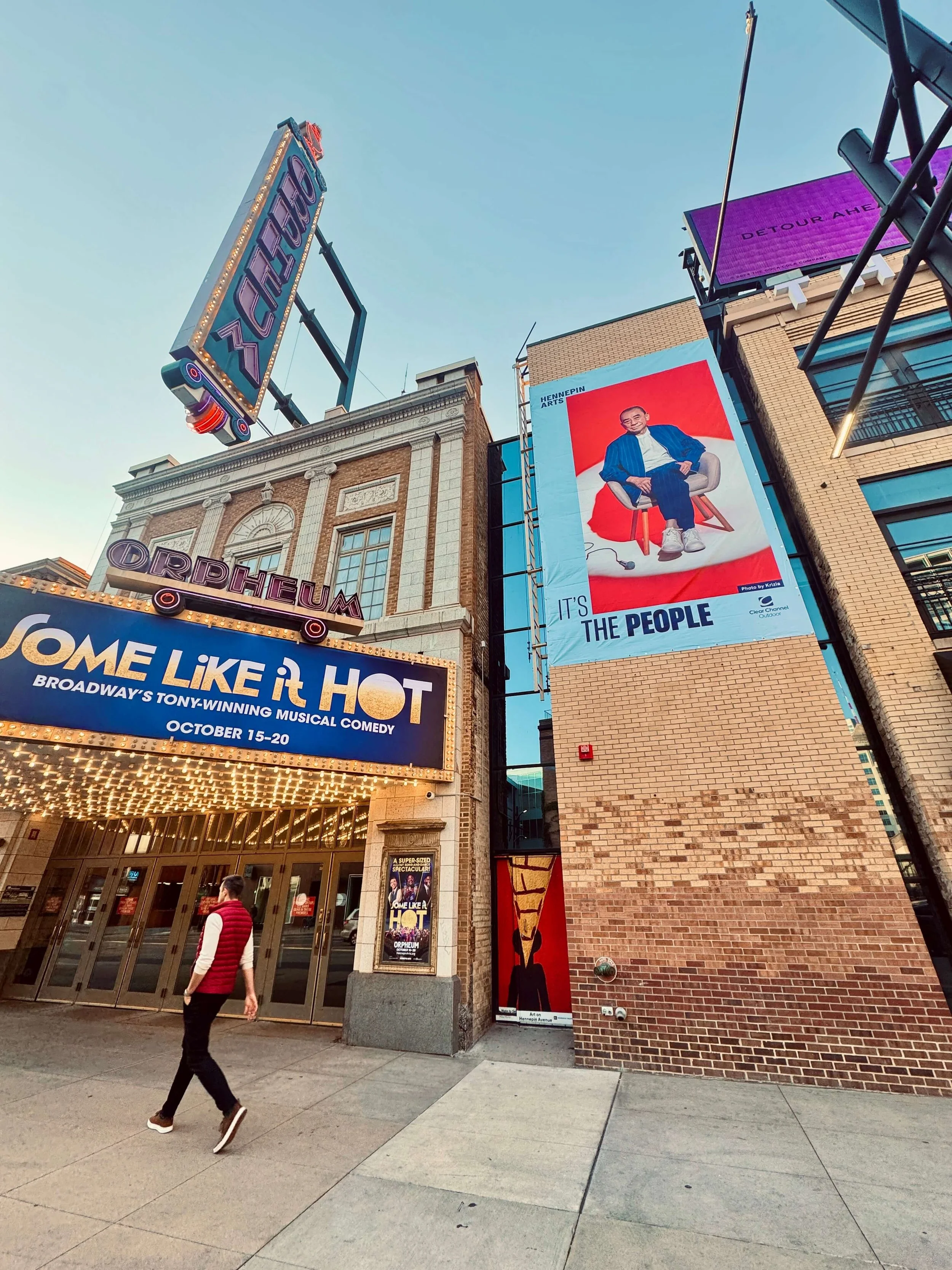 The Orpheum Theatre entrance with marquee promoting Broadway's 'Come Like It HOT' musical, a large billboard with an artwork of a man sitting in a chair labeled 'It's the People', and surrounding signage and posters.