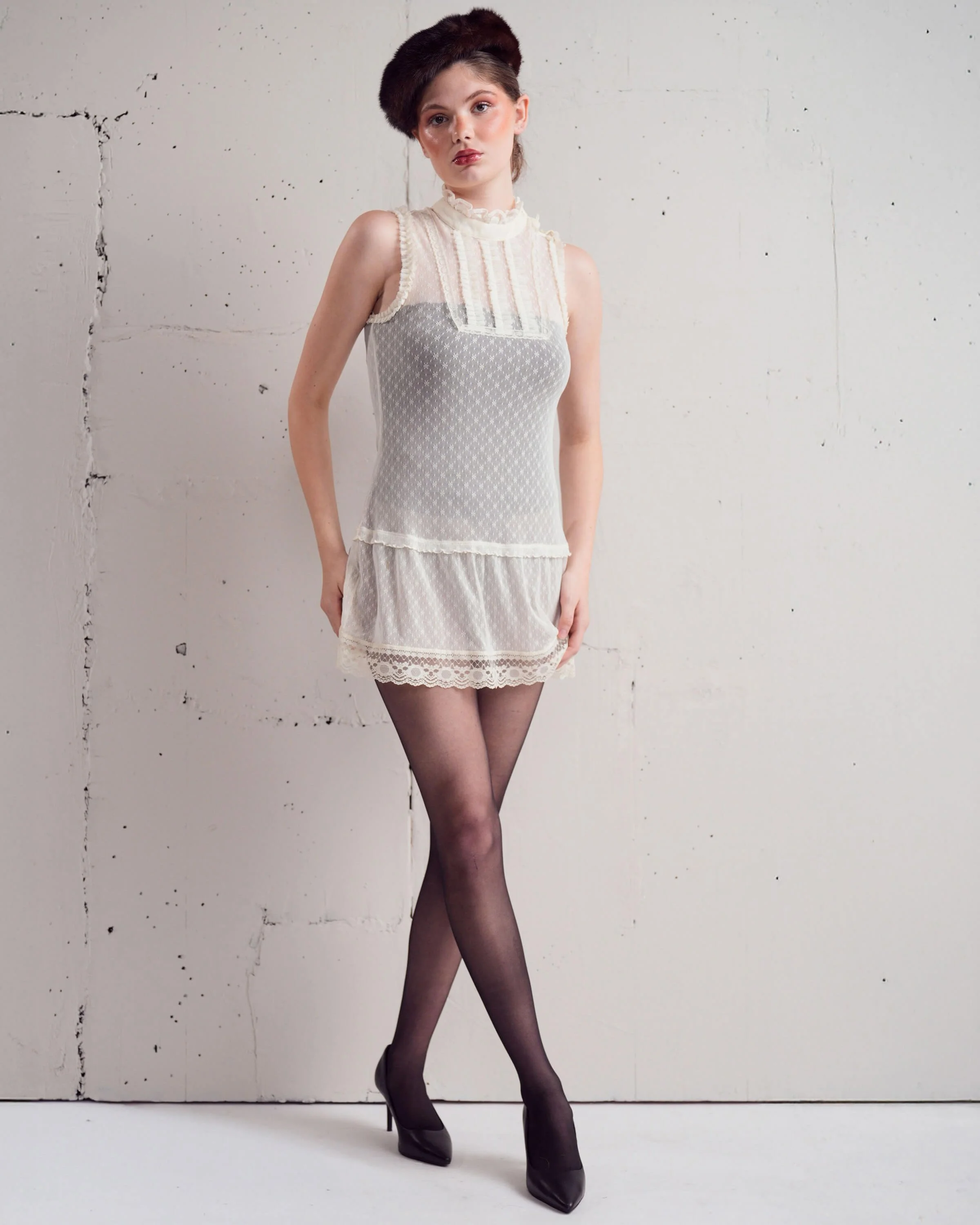 Young woman with dark hair styled up, wearing a vintage-inspired beige dress with lace details, black sheer tights, pointed black heels, standing against a plain white wall with some visible cracks.