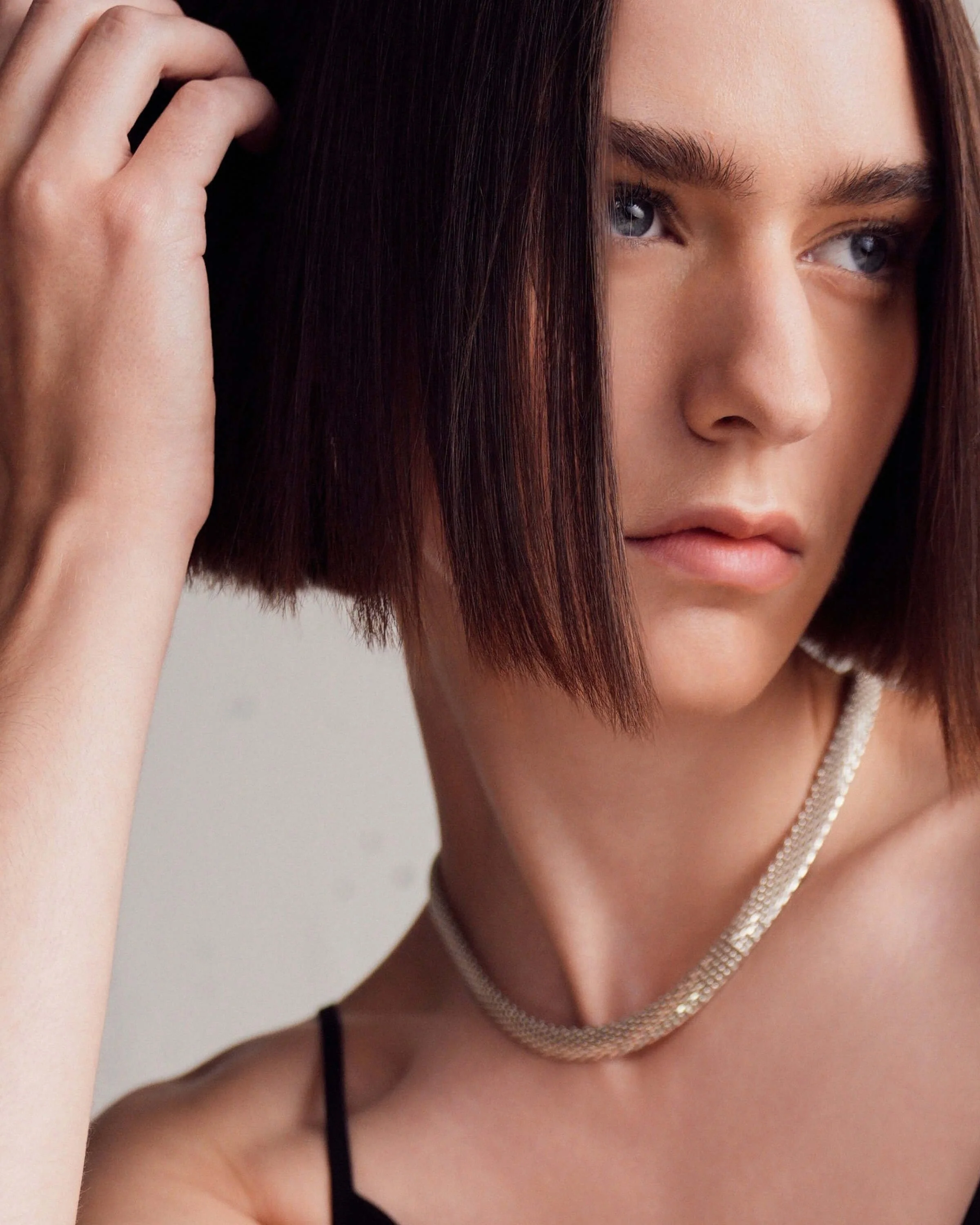 Close-up of a young woman with shoulder-length brown hair, blue eyes, and fair skin. She is wearing a silver necklace and a black spaghetti strap top, with her hand touching her hair.