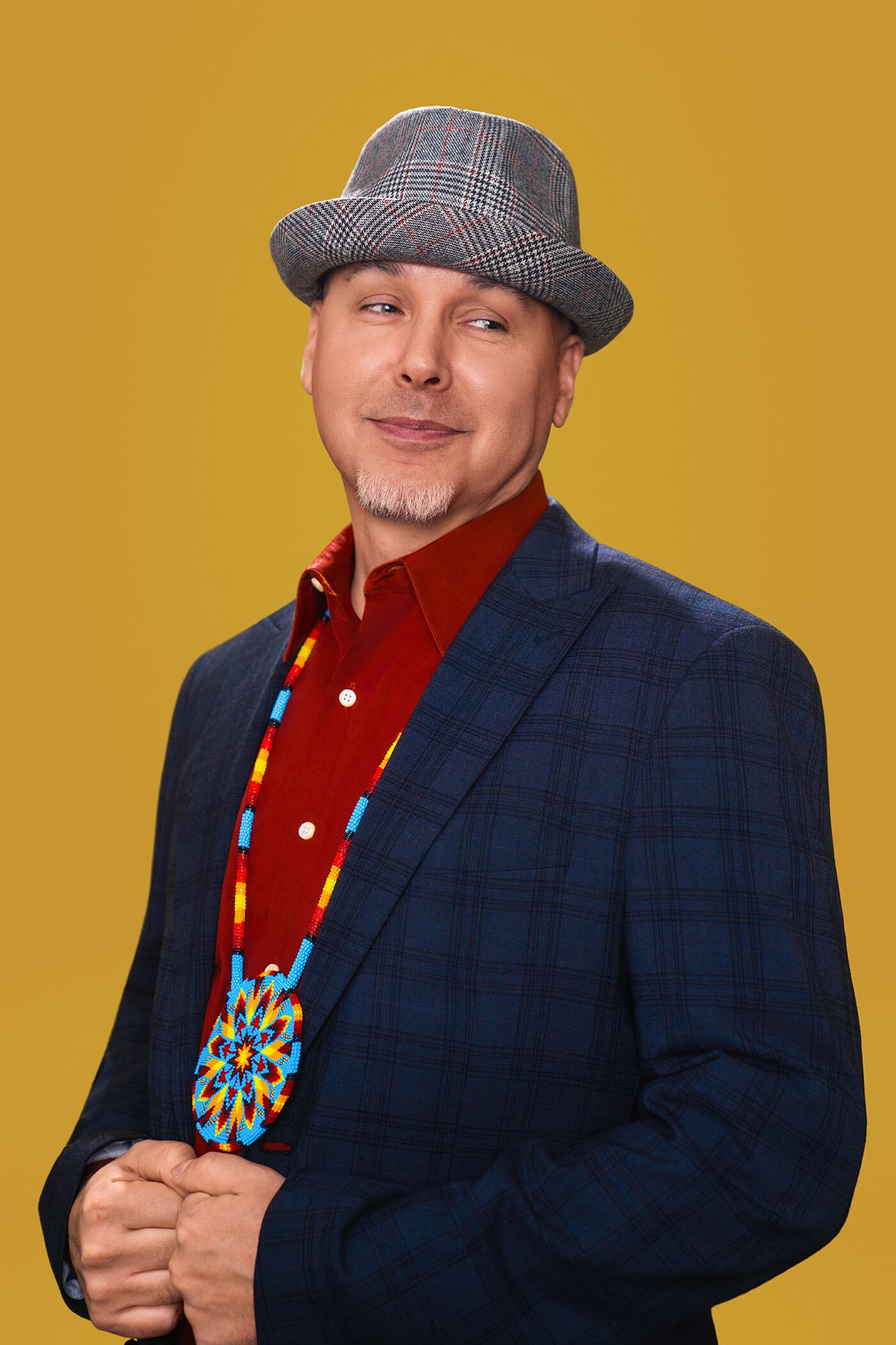 A man wearing a gray plaid fedora, a red shirt, a navy blazer, and a beaded necklace with a Native American design, standing against a yellow background.