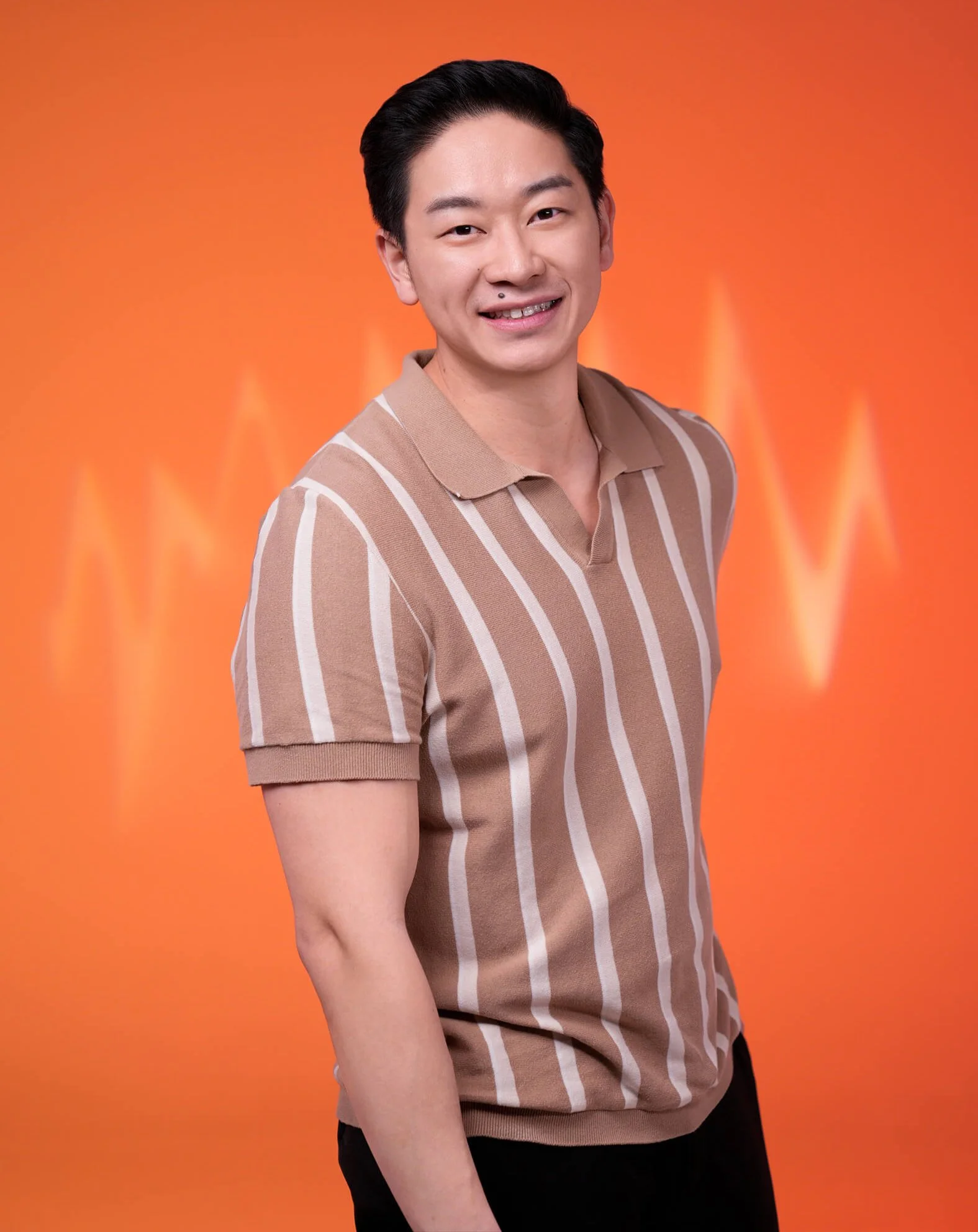 A smiling young man with dark hair wearing a tan and white striped polo shirt, standing against an orange background with a subtle heartbeat line graphic.