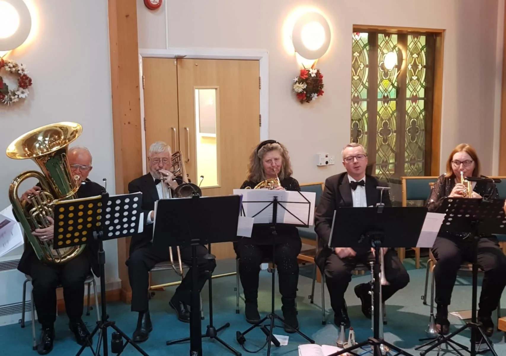 A jazz band performing indoors with five musicians playing various brass instruments, seated behind music stands.