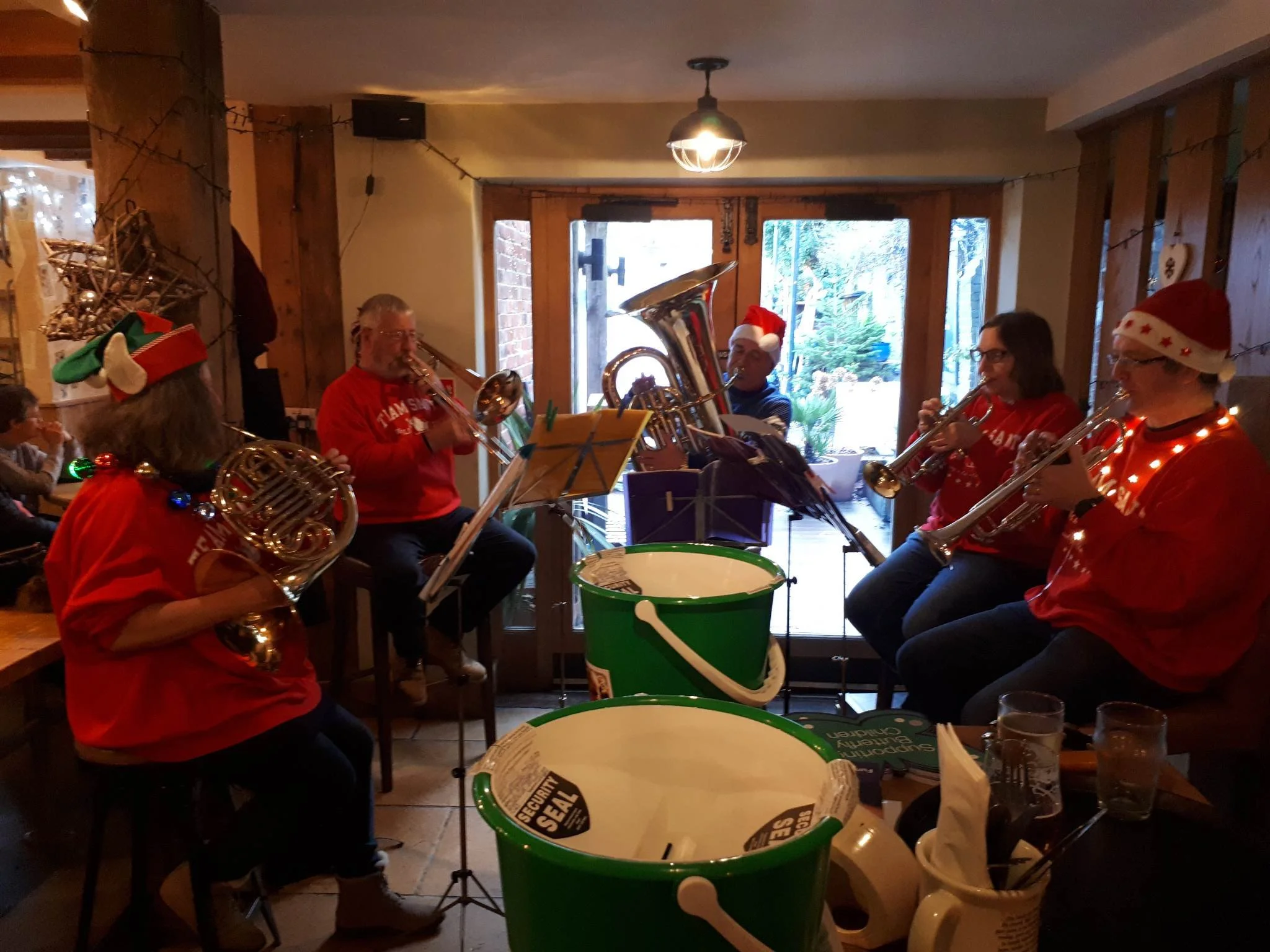 A group of people playing brass instruments in a cozy room with wood paneling, Christmas hats and sweaters, and holiday decorations, during a festive gathering.