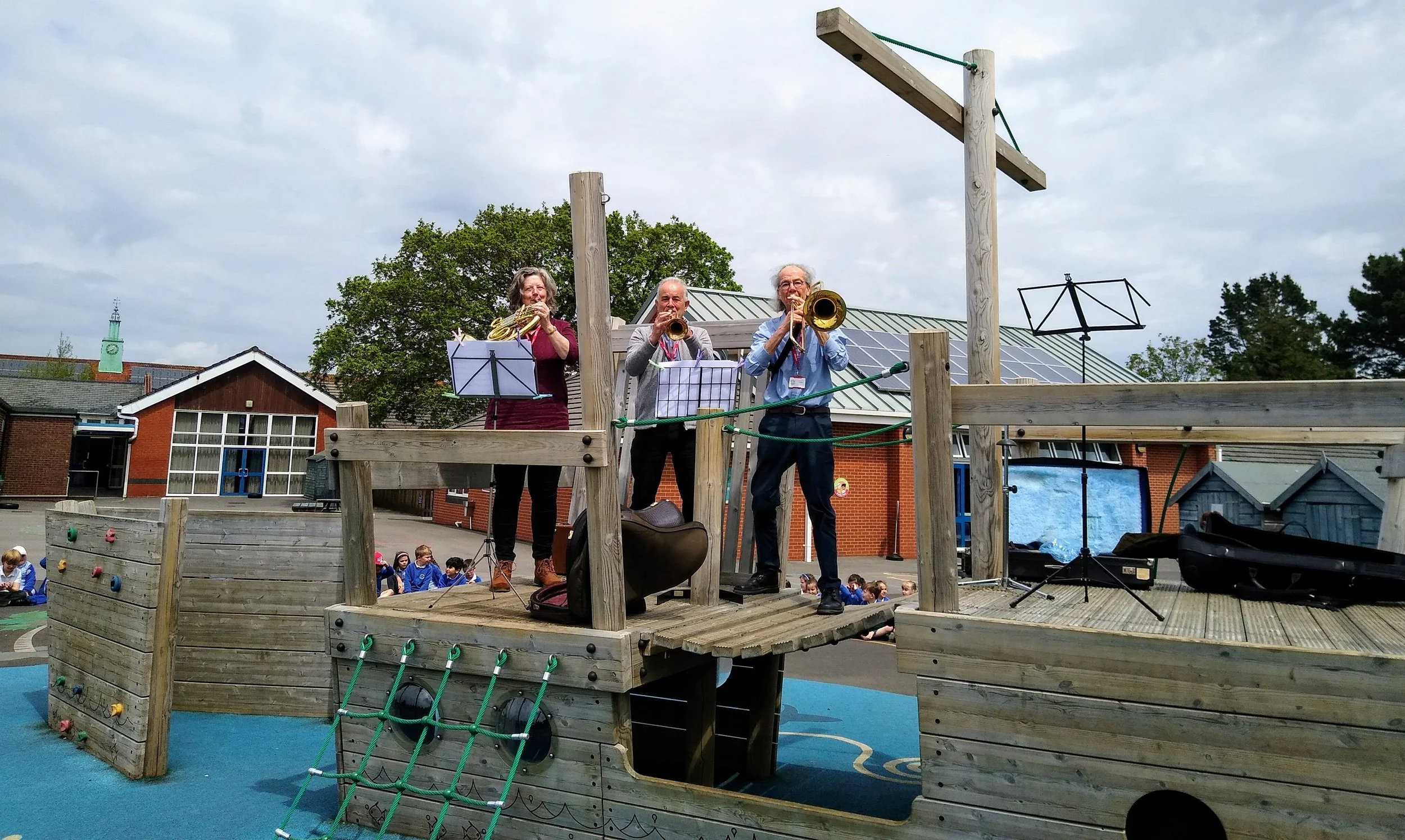 Three musicians playing wind instruments on a wooden playground structure outdoors, with children sitting on the ground in the background.