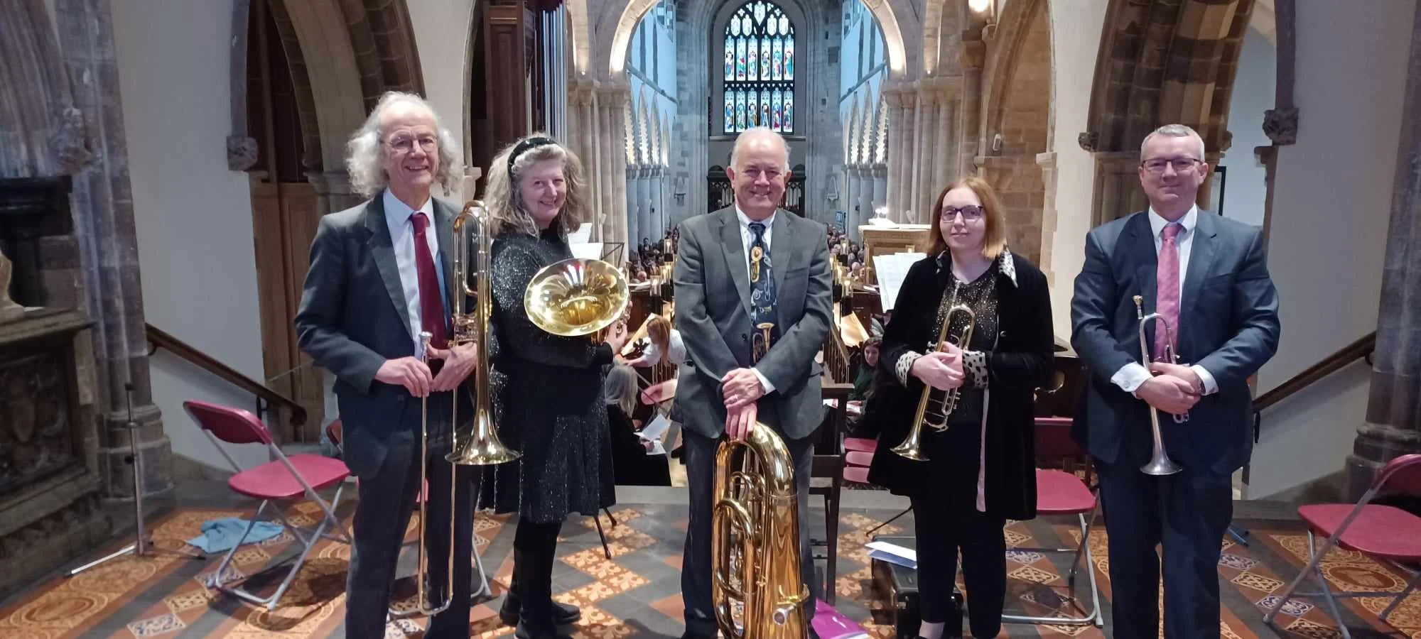 Five musicians with brass instruments standing inside a church with stained glass windows and an audience behind them.