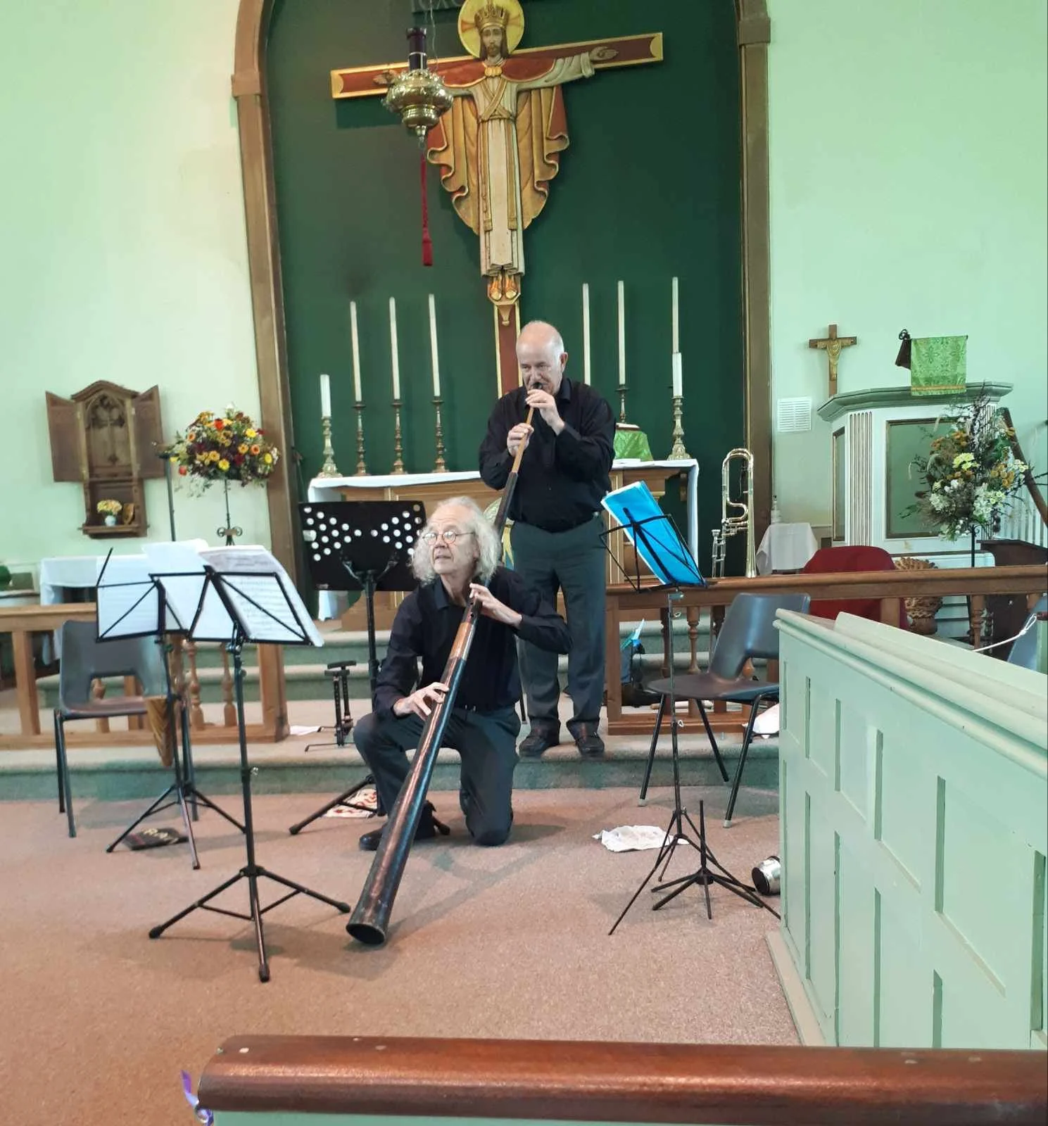 Two musicians performing with wind instruments inside a church with a crucifix, candles, and floral arrangements in the background.
