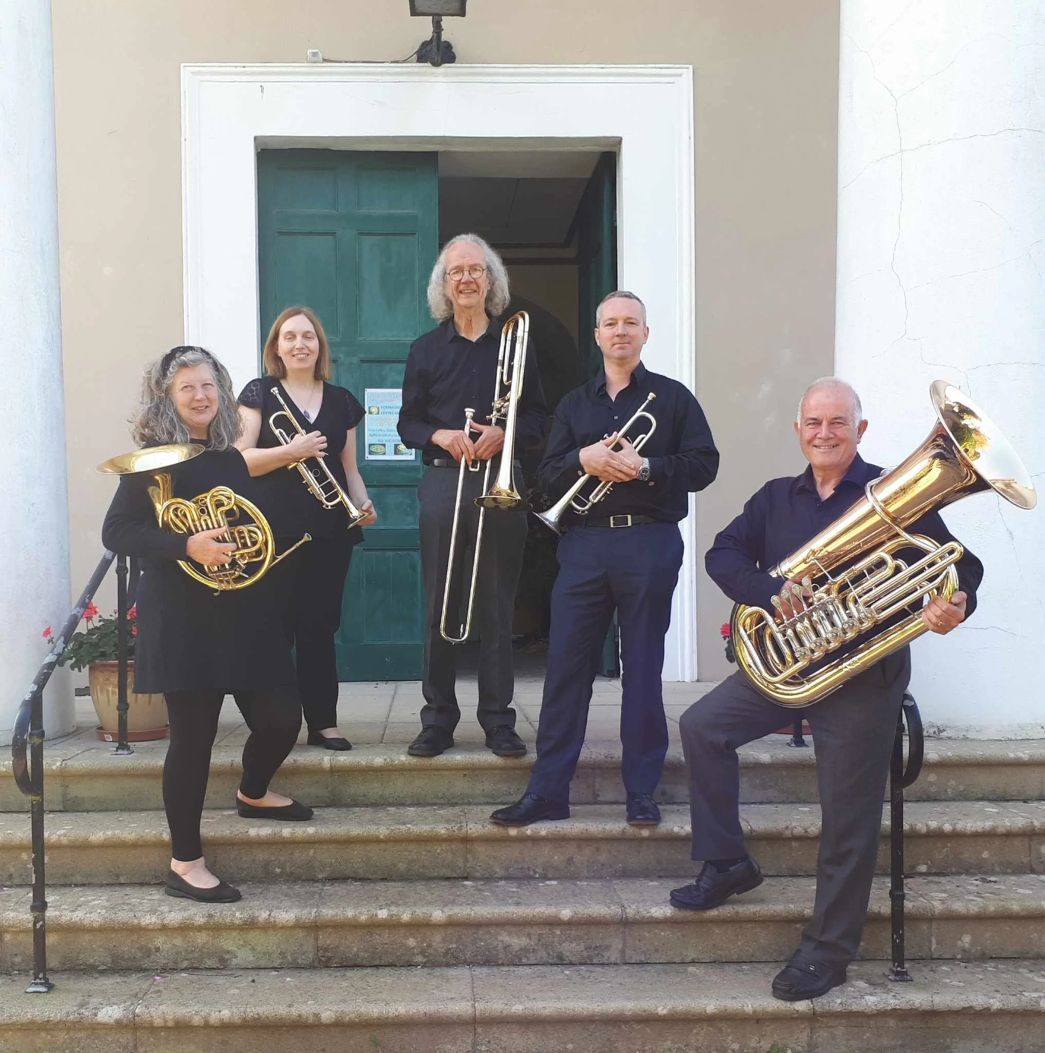 A group of five musicians with brass instruments standing on the steps of a building with a green door, smiling at the camera.