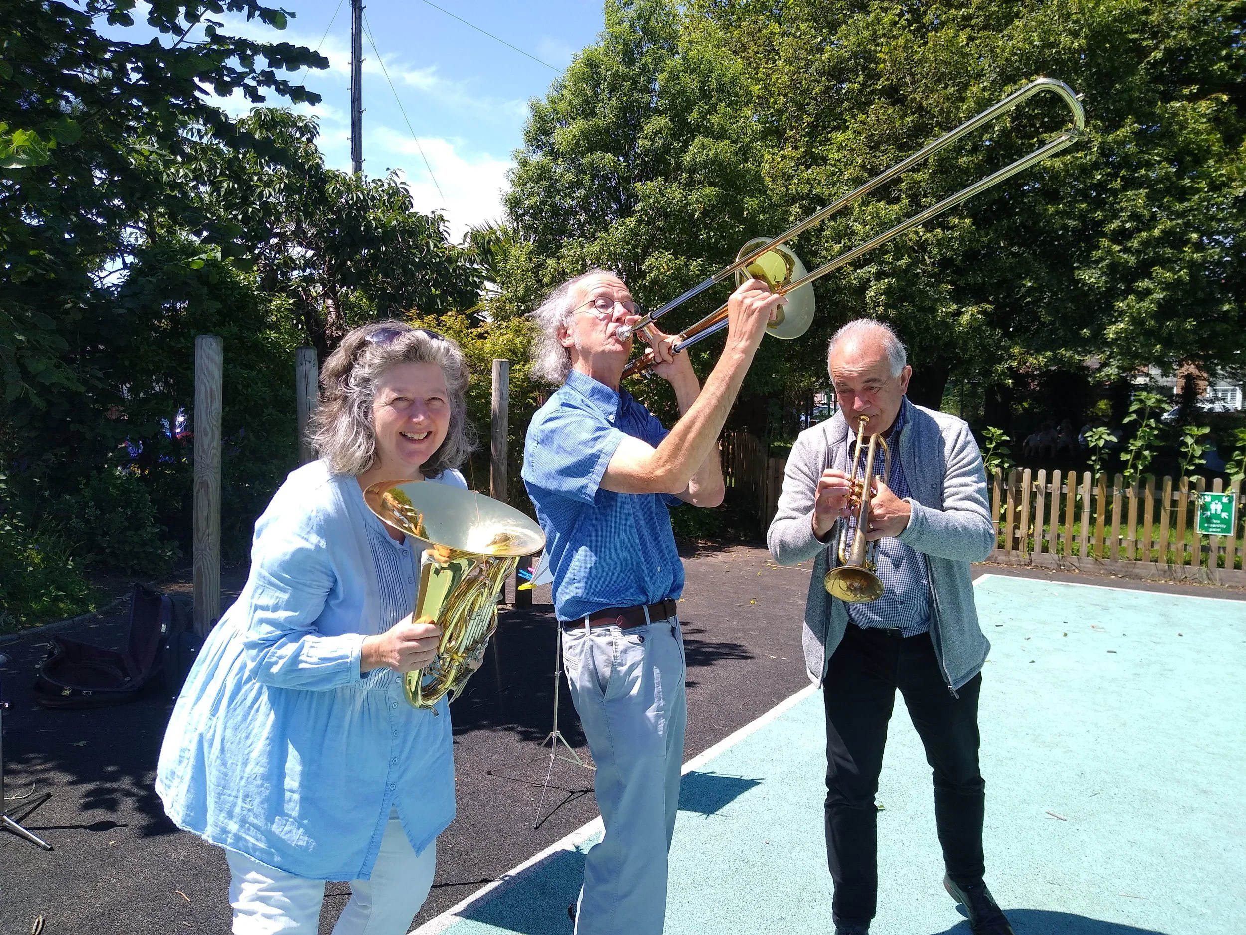 Three older adults playing musical instruments outdoors on a sunny day. The woman on the left is holding a brass instrument, the man in the middle is playing a trombone, and the man on the right is playing a trumpet. They are standing on a sports court with trees and a wooden fence in the background.