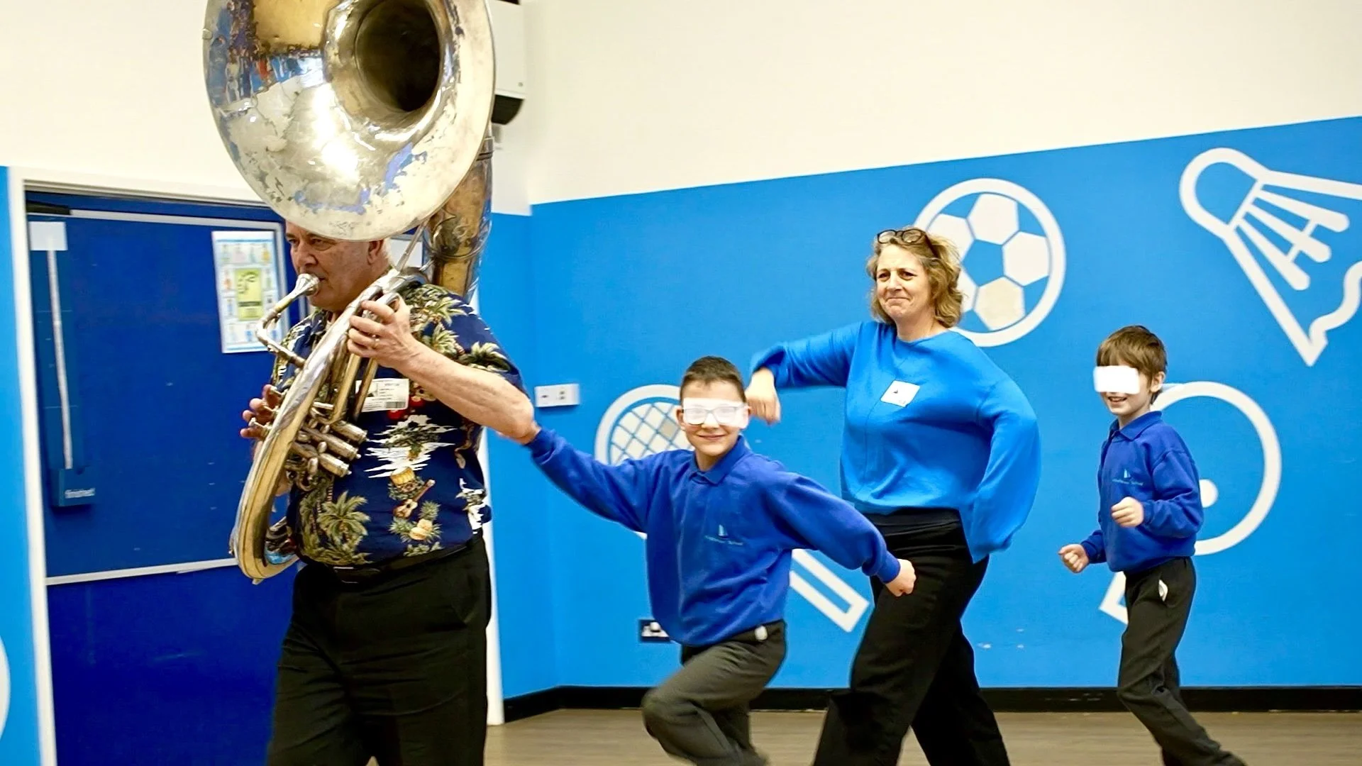 An elderly man playing a sousaphone, with a woman and two children dancing behind him in a room with blue sports-themed wall decor.