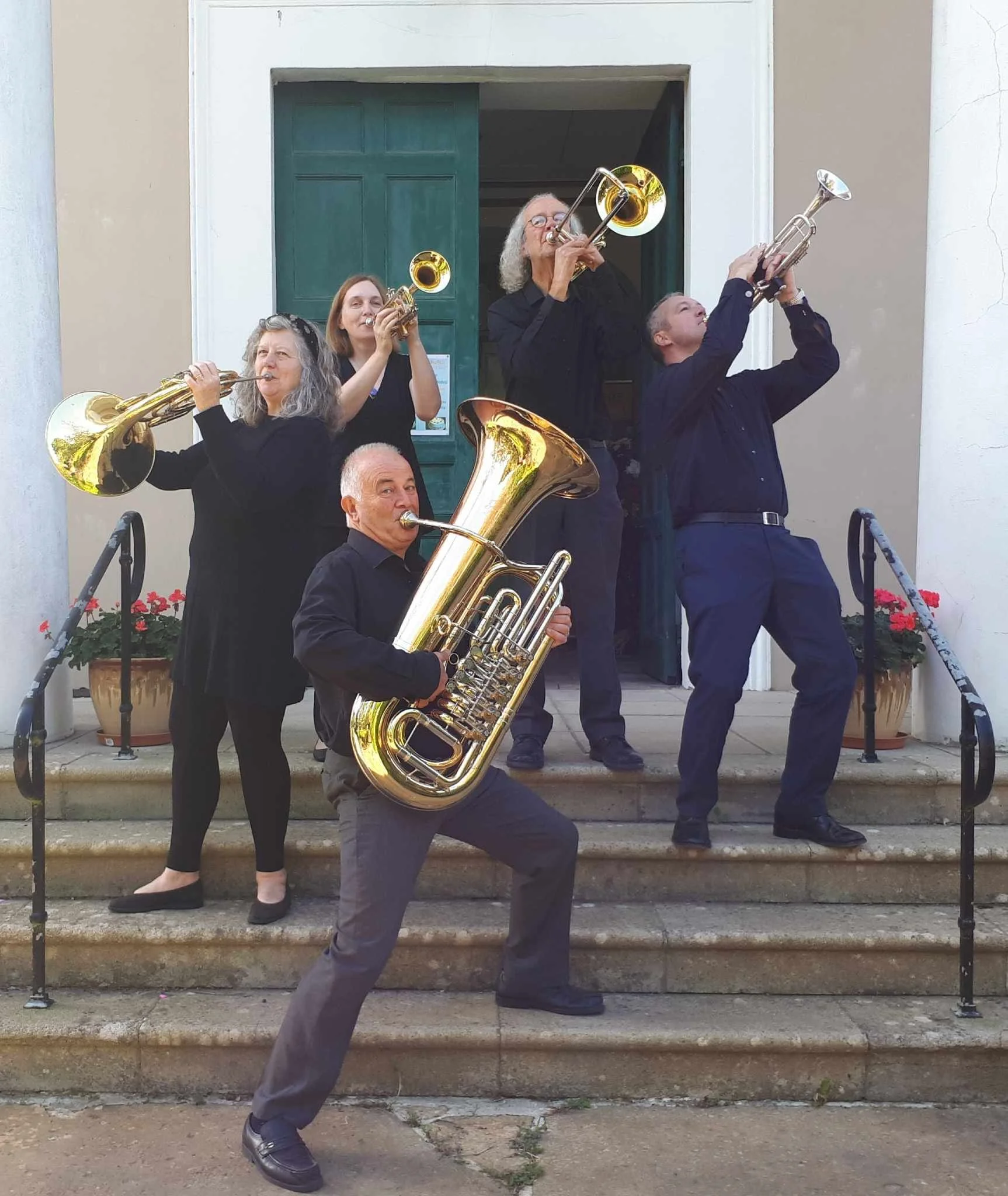 Group of five musicians playing brass instruments on steps outside a building with green doors, including a man with a tuba, a woman with a trumpet, an older man with a trumpet, and two women with trumpets.