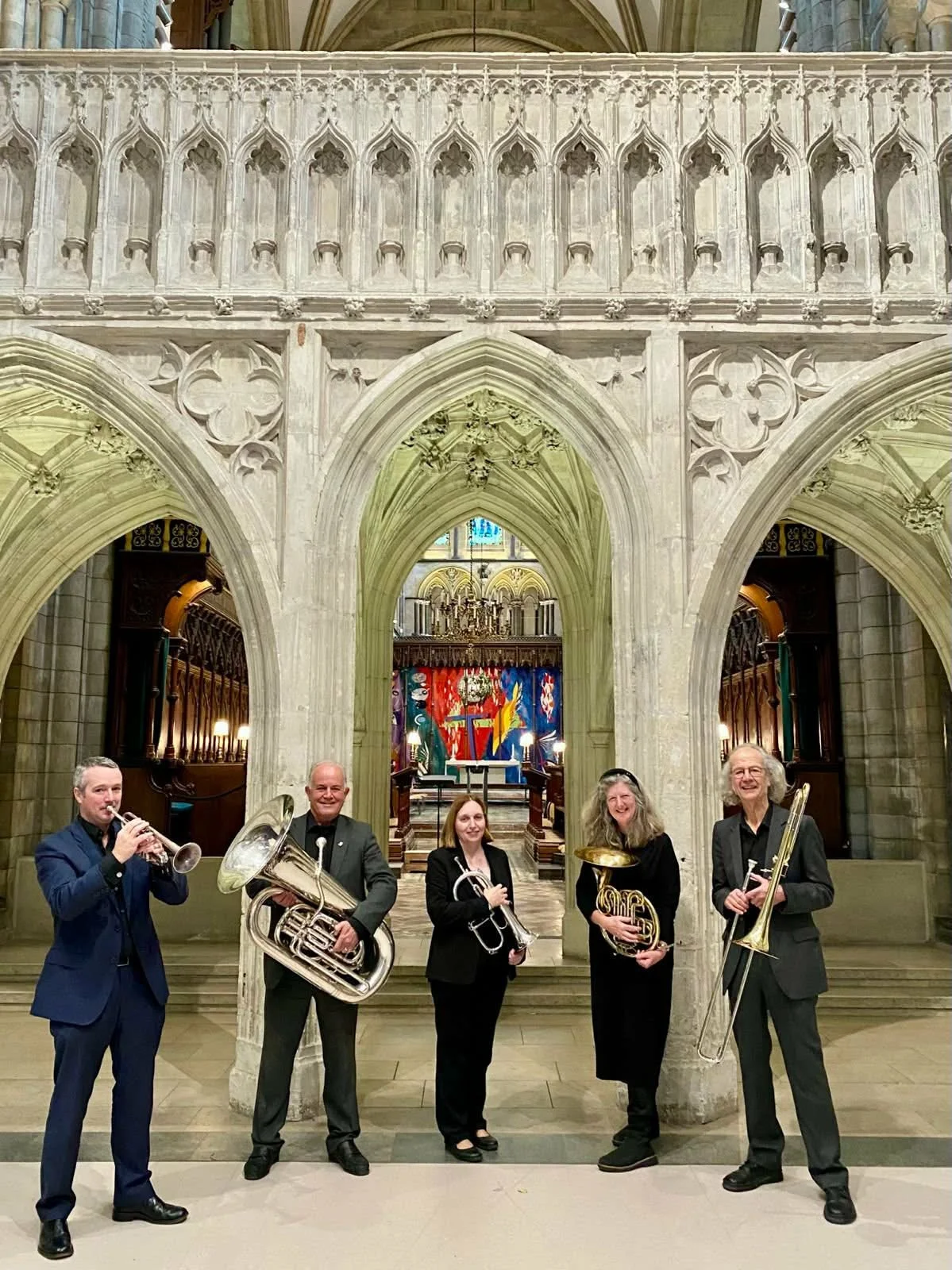 Five musicians holding brass instruments standing inside a Gothic-style church or cathedral with stone arches, intricate architecture, and religious artwork in the background.