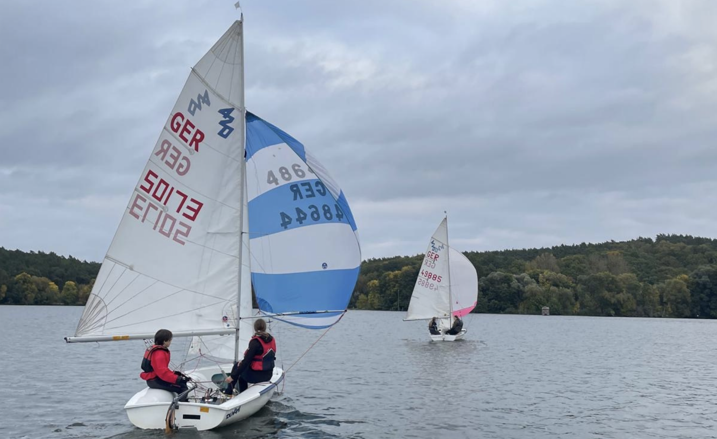 Segelboote auf einem See mit Menschen an Bord und städtisches Hinterland mit Bäumen, im September bei bewölktem Himmel.
