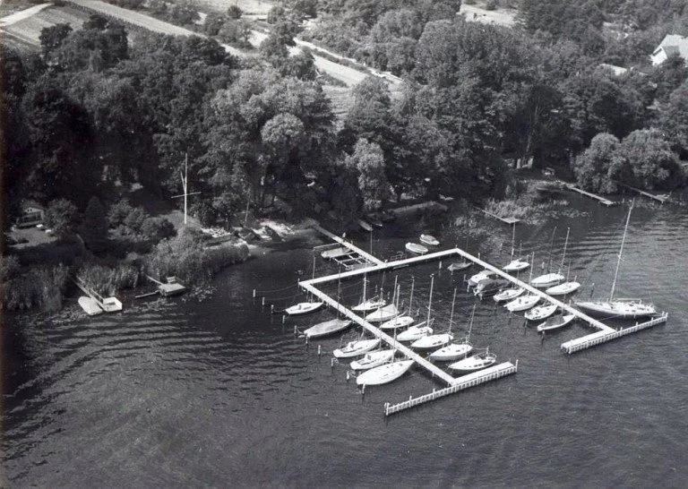 Luftaufnahme eines Yachthafens mit mehreren anliegenden Booten, umgeben von Bäumen und einem Gewässer, mit einer ruhigen, bewaldeten Uferlandschaft im Hintergrund.