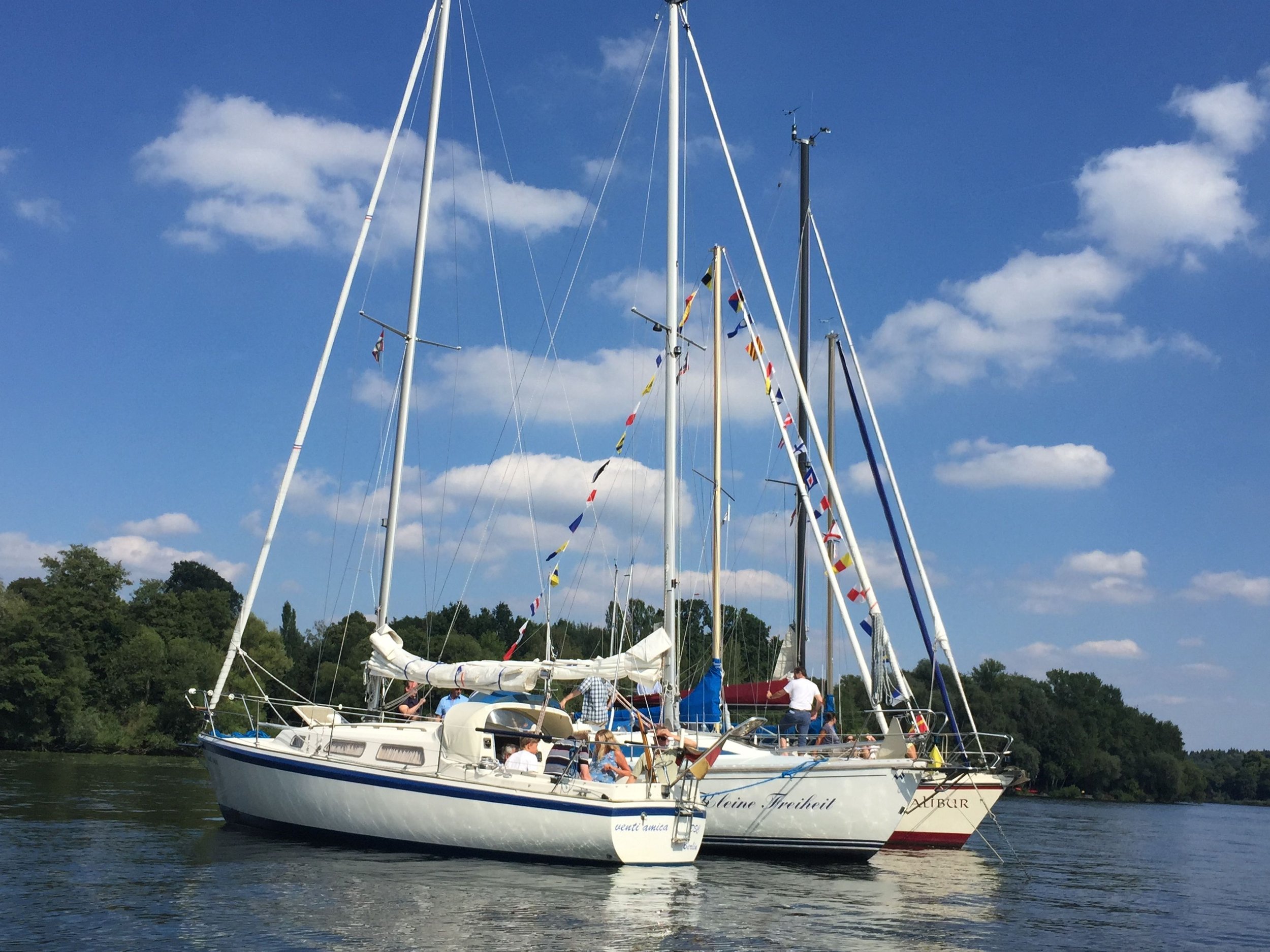 Ein weißes Segelboot mit mehreren Menschen an Bord liegt an einem Fluss oder See, bei sonnigem Himmel mit einigen Wolken, im Hintergrund Bäume.