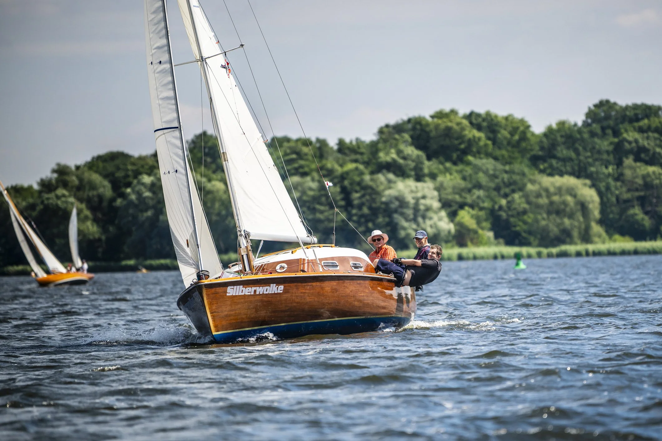 Menschen segeln auf einem Holzboot namens Silberwolke auf einem See mit grünem Baumbestand im Hintergrund.