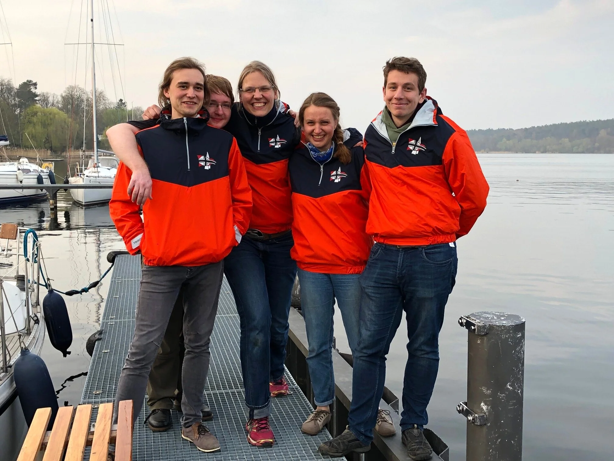 Fünf junge Menschen, alle tragen orange- und dunkelblaue Jacken mit einem Kreuz-Logo, posieren auf einem Steg am Wasser mit Booten im Hintergrund, wahrscheinlich nach einem Segel- oder Bootswettbewerb.
