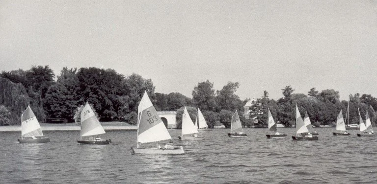 Mehrere kleine Segelboote auf einem See bei ruhigem Wasser, mit Bäumen im Hintergrund.