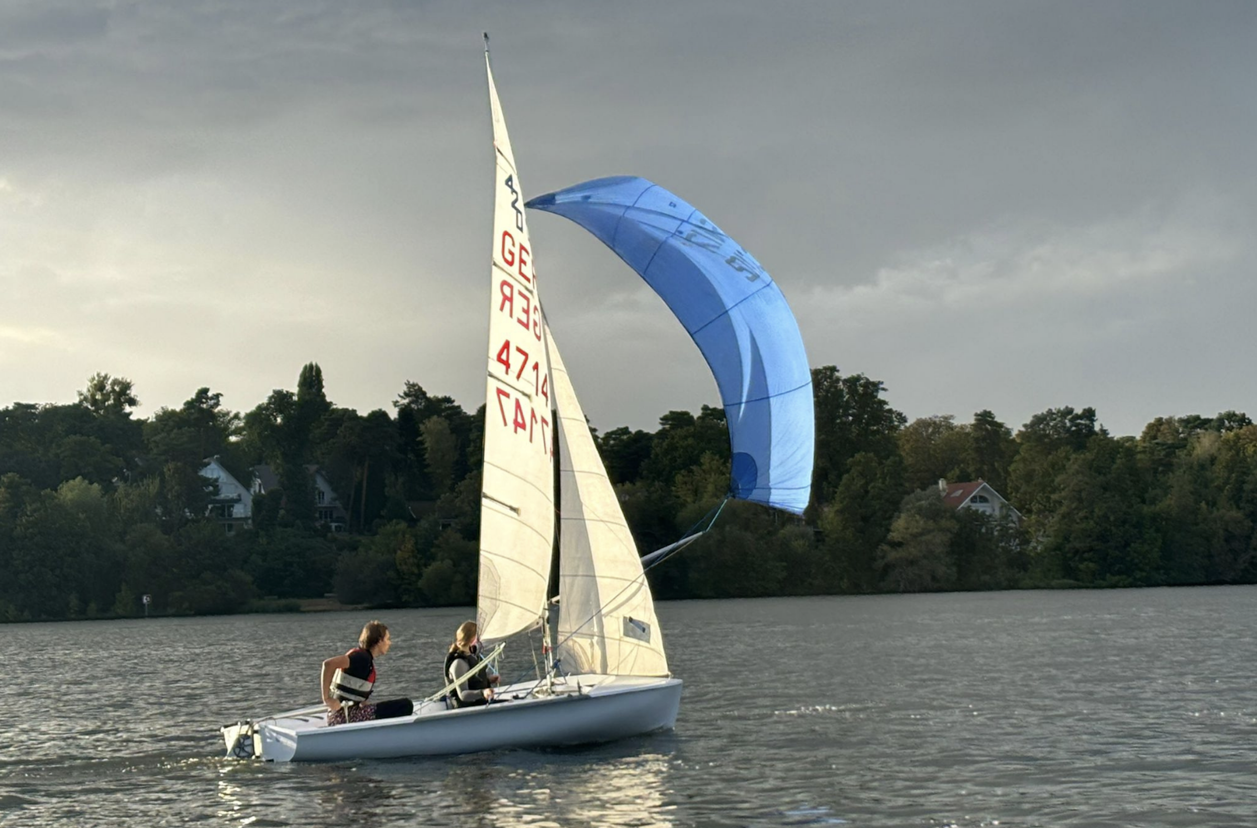 Zwei Frauen segeln auf einem kleinen Boot auf dem Wasser, mit Wolken im Himmel und Häusern sowie Bäumen im Hintergrund.
