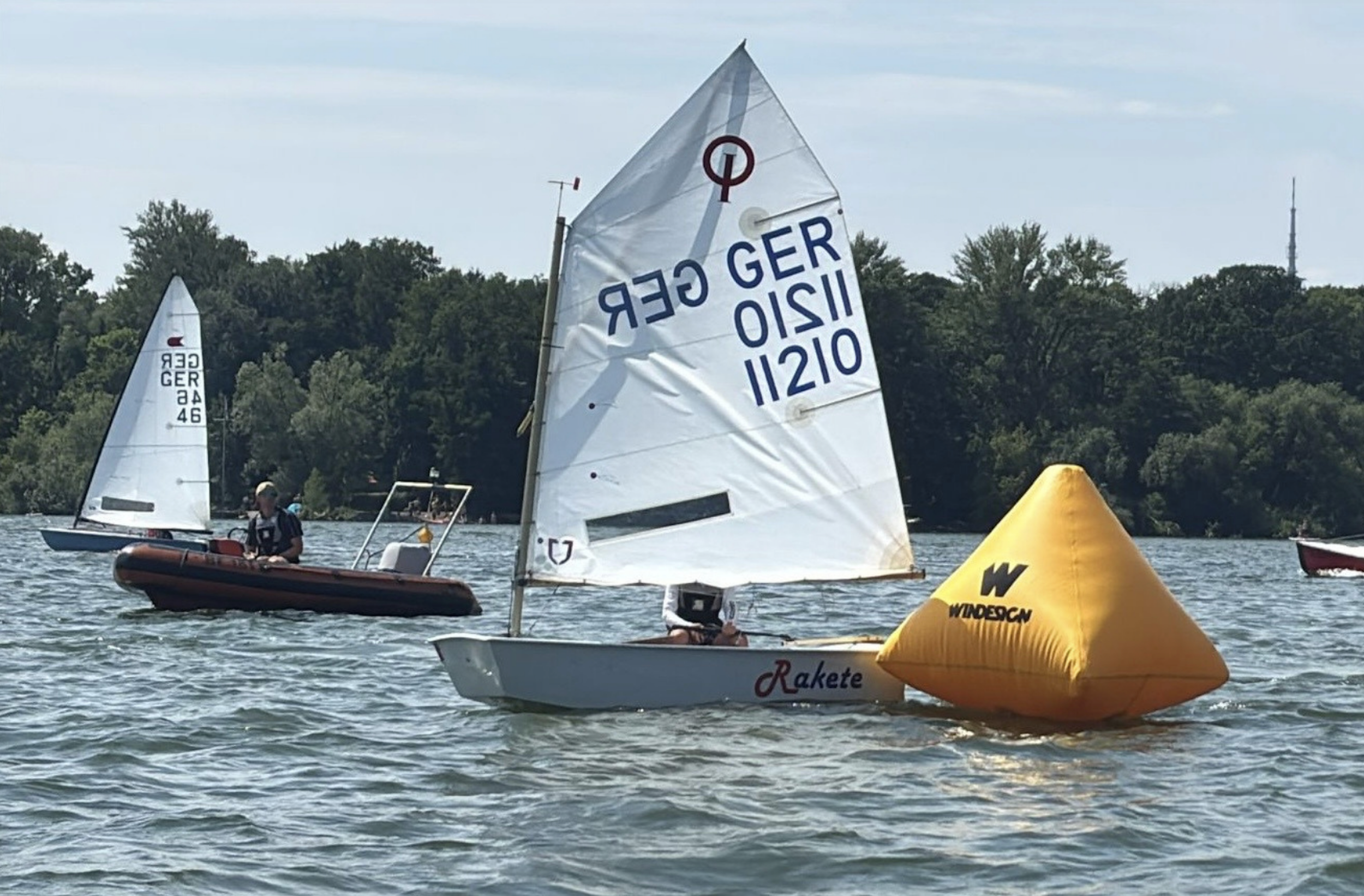Segelboote auf dem Wasser während eines Wettkampfs, mit einer gelben Windfahne im Vordergrund, im Hintergrund Bäume und ein Fernsehturm.