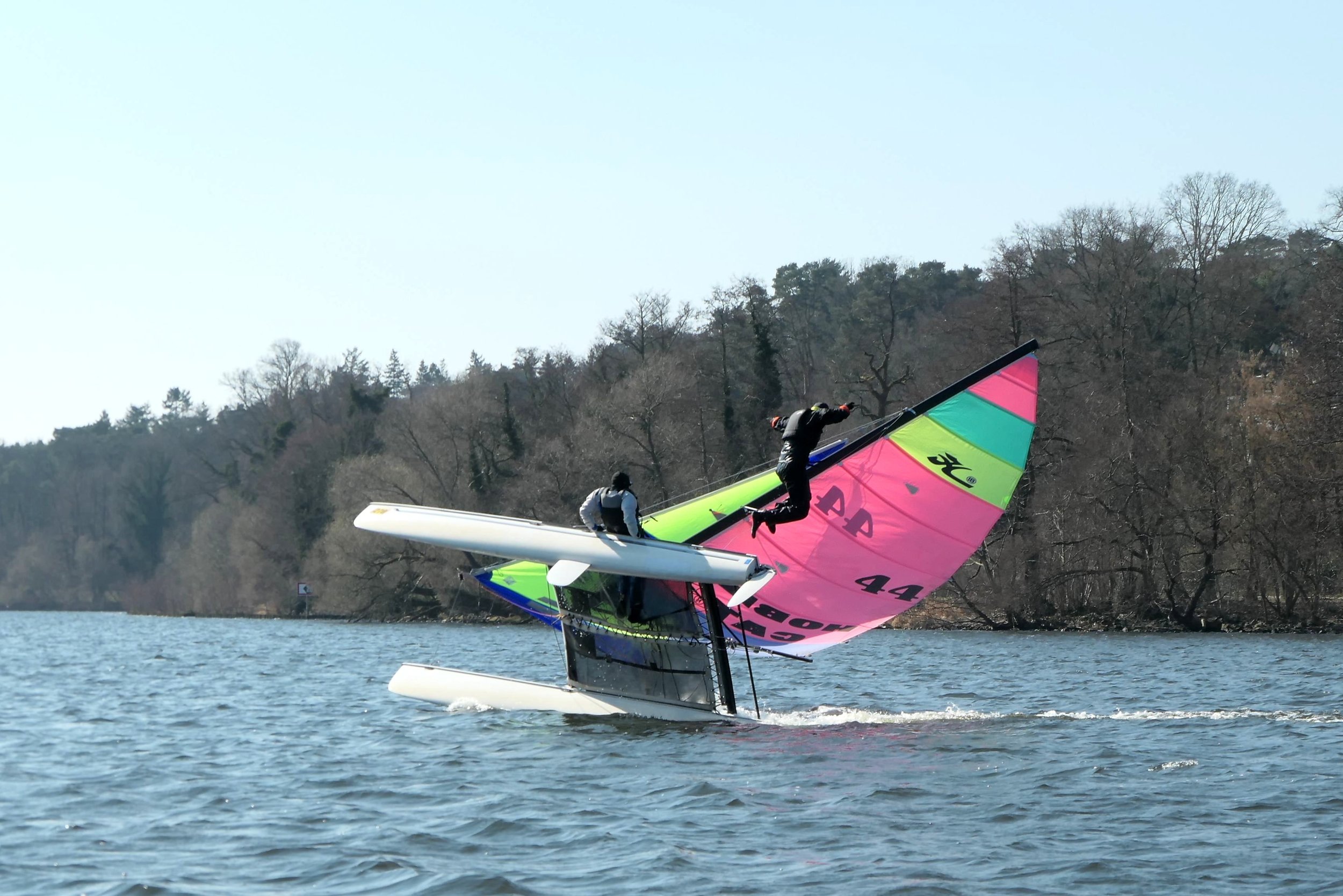 Segler auf einem Wasserfahrzeug mit buntem Segel, das über das Wasser gleitet, mit einem Hintergrund aus Bäumen und Himmel.