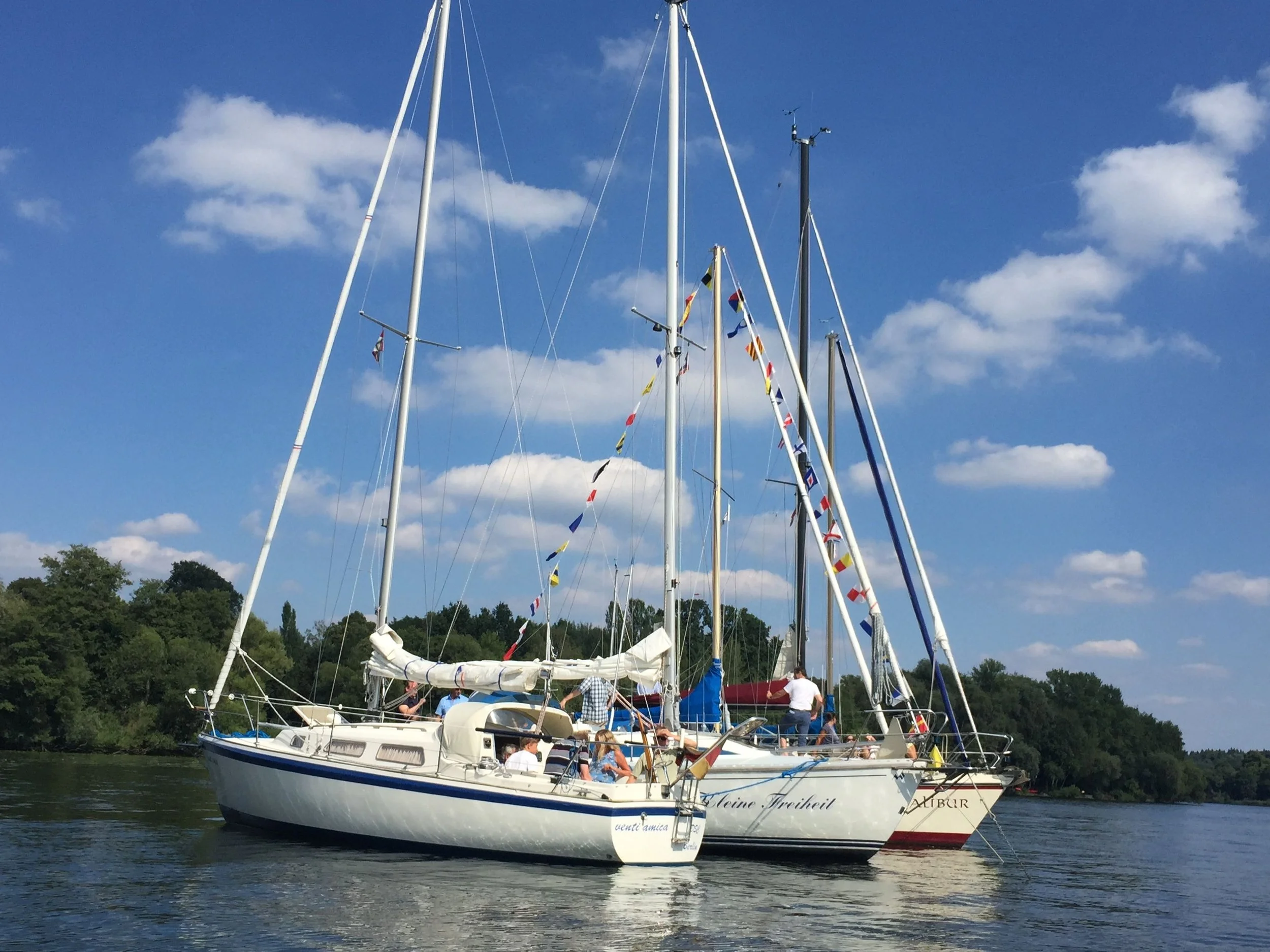 Segelboot mit Flaggen am Wasser, Menschen an Bord, im Hintergrund Bäume und ein blauer Himmel mit Wolken.