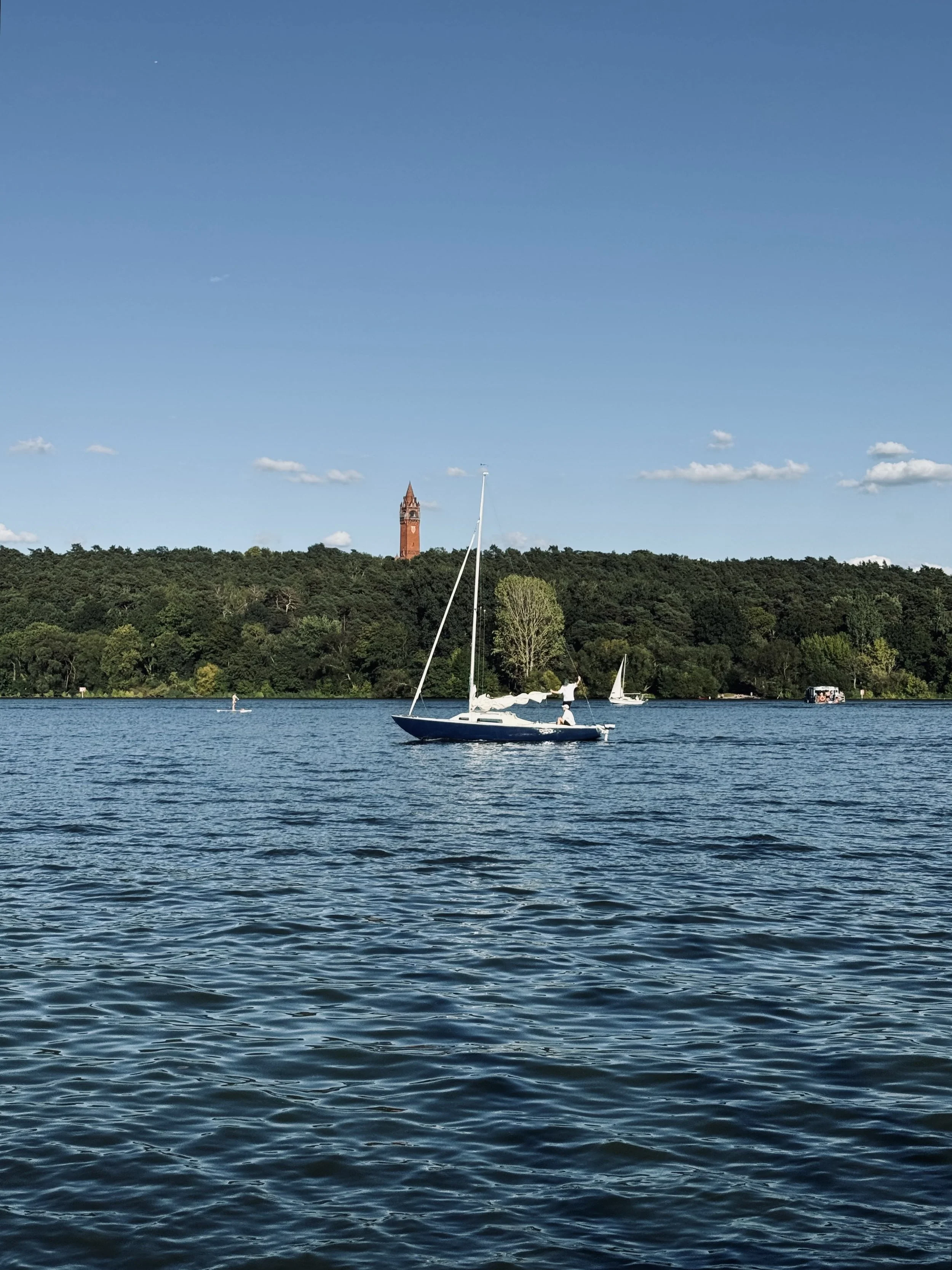Unterer Teil eines Sees mit Wasser, im Hintergrund bewaldete Hügel, eine weiße Segelyacht und zwei kleinere Boote. Über dem Himmel befinden sich wenige Wolken, in der Ferne erkennt man einen Kirchturm.
