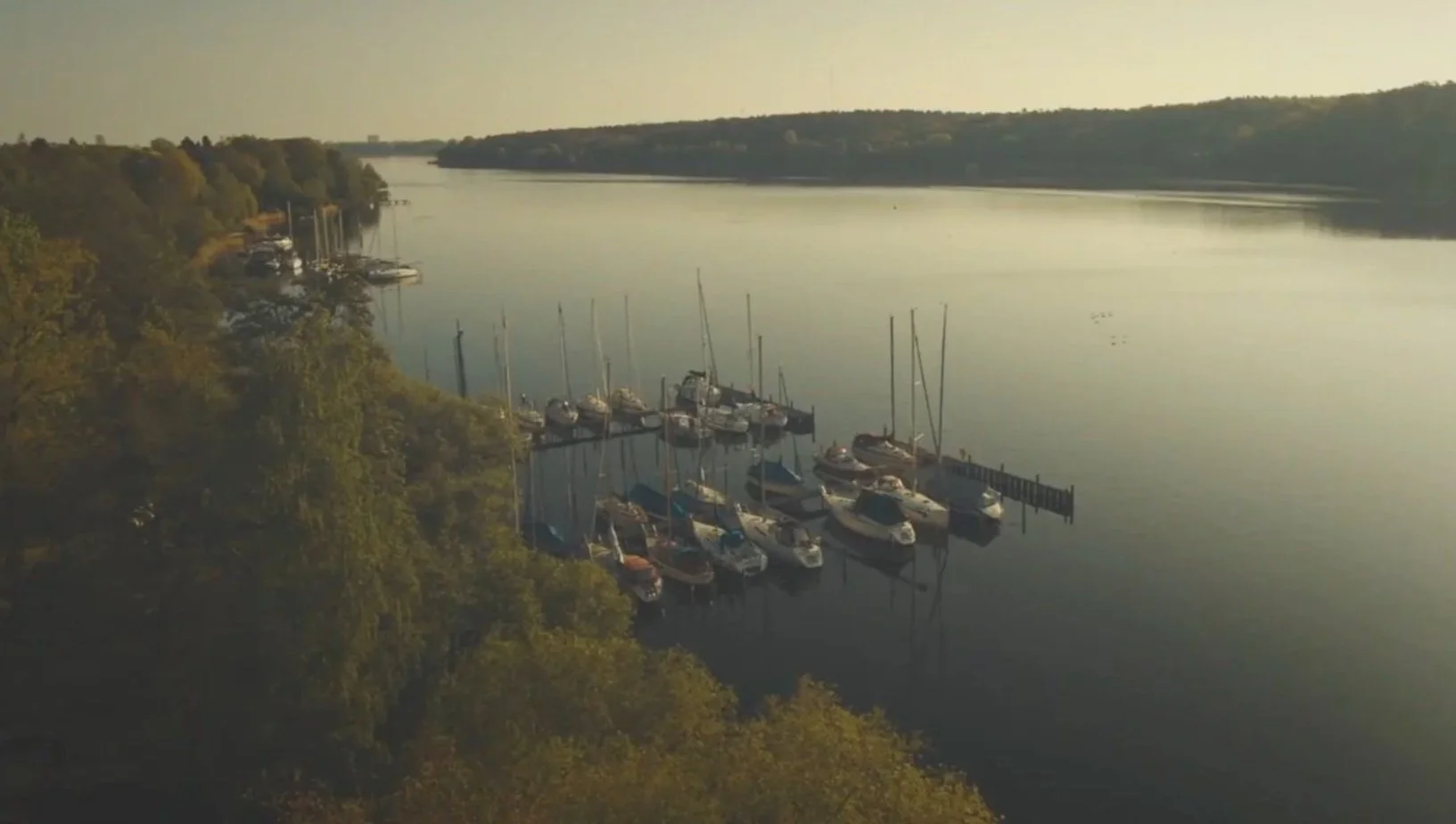 Der Blick auf einen ruhigen Fluss mit ankerten Booten und Segelbooten an einem Dock, umgeben von dichter grüner Vegetation und sanften Hügeln im Hintergrund bei Sonnenuntergang.