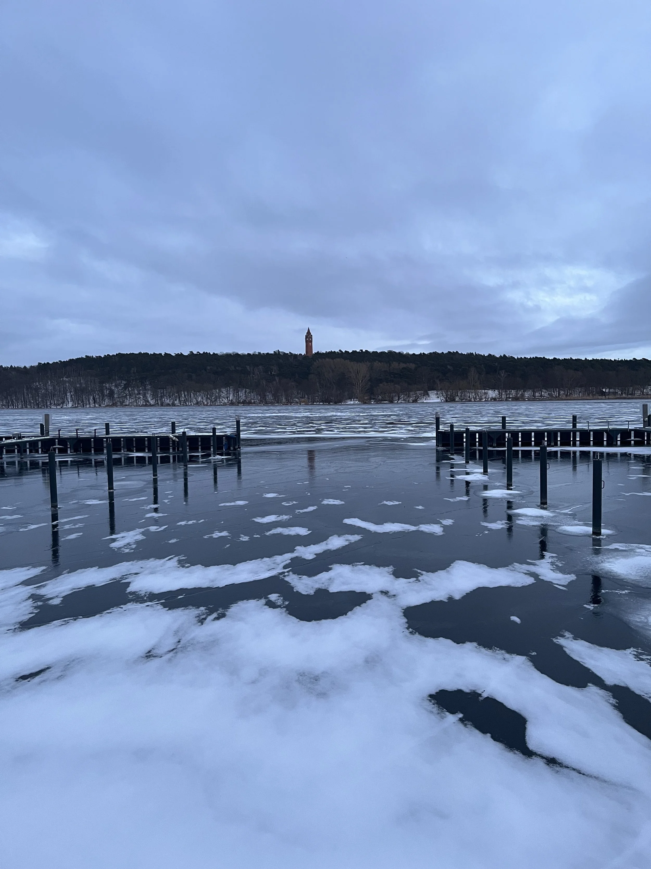 Der Himmel ist bewölkt, und das Wasser ist teilweise zugefroren, mit Eisschollen und Schneeflächen. Im Hintergrund ist ein Hügel mit Bäumen, und auf der Spitze des Hügels steht ein Turm.
