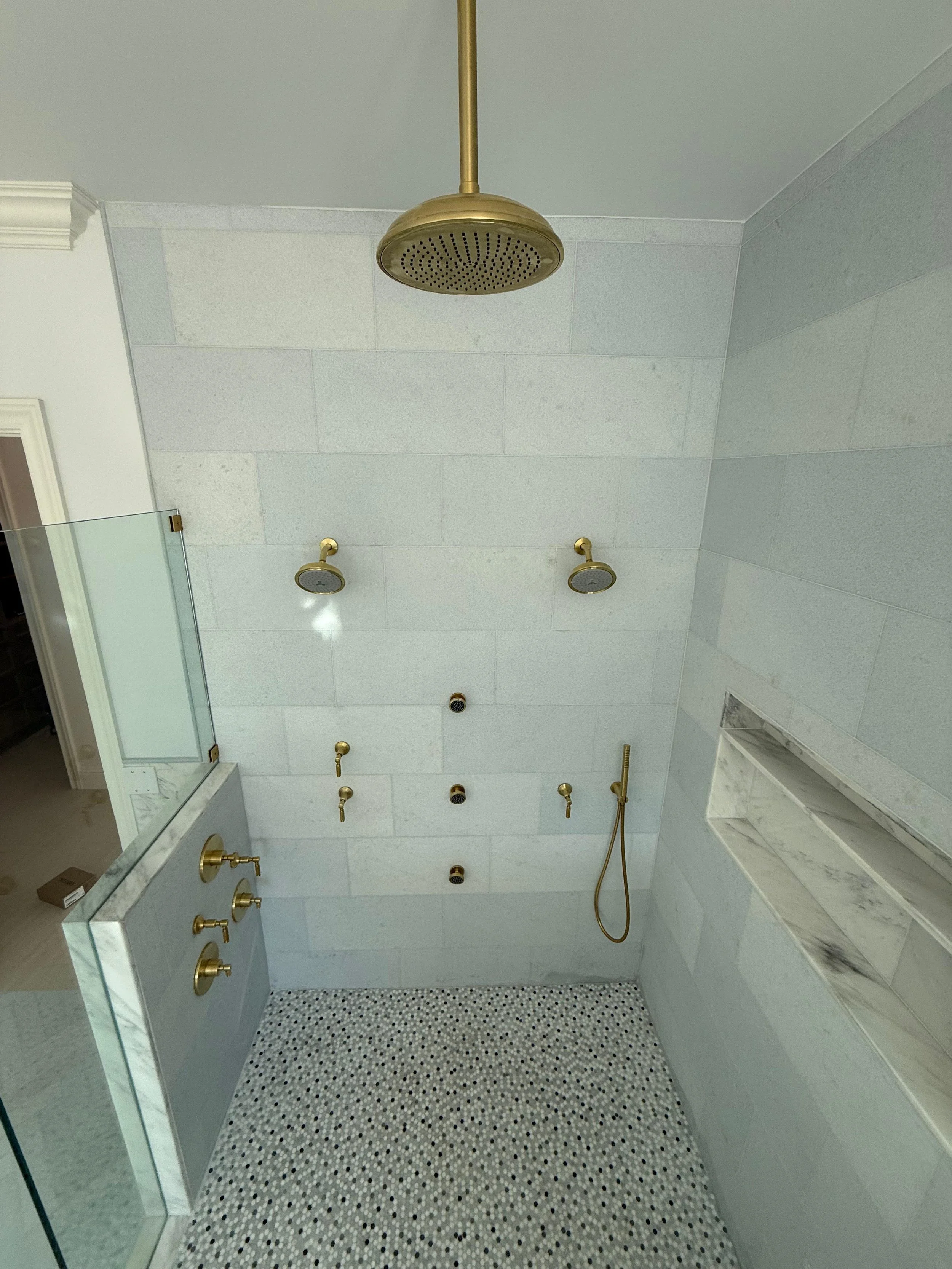 Empty walk-in shower with a gold rain shower head, two smaller shower heads, a handheld shower, and a marble shelf with a gray and white pattern. The shower has white stone tiles and a small mosaic tile floor.