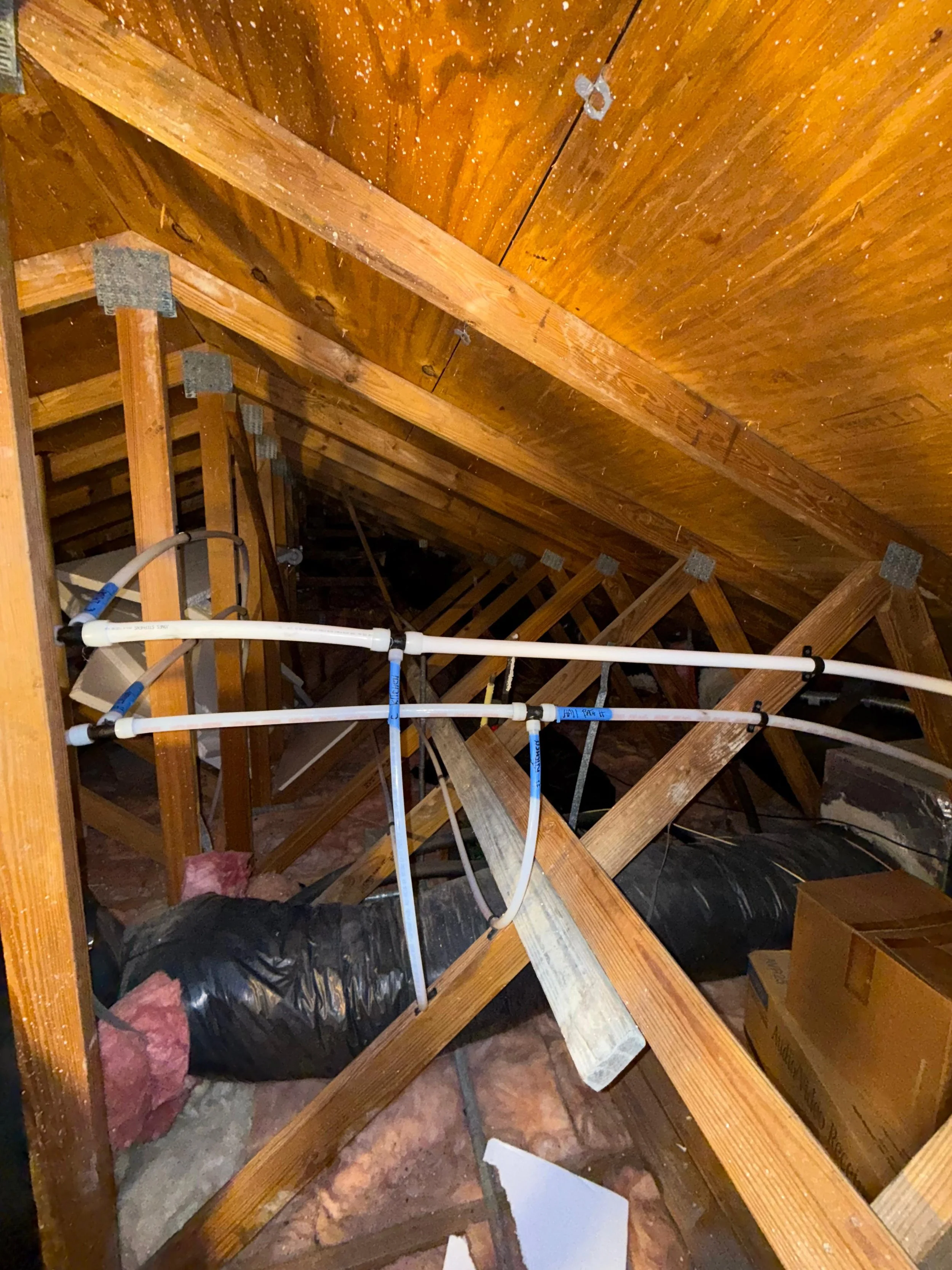 Attic with exposed wooden beams, PVC pipe plumbing, insulation, and a cardboard box.