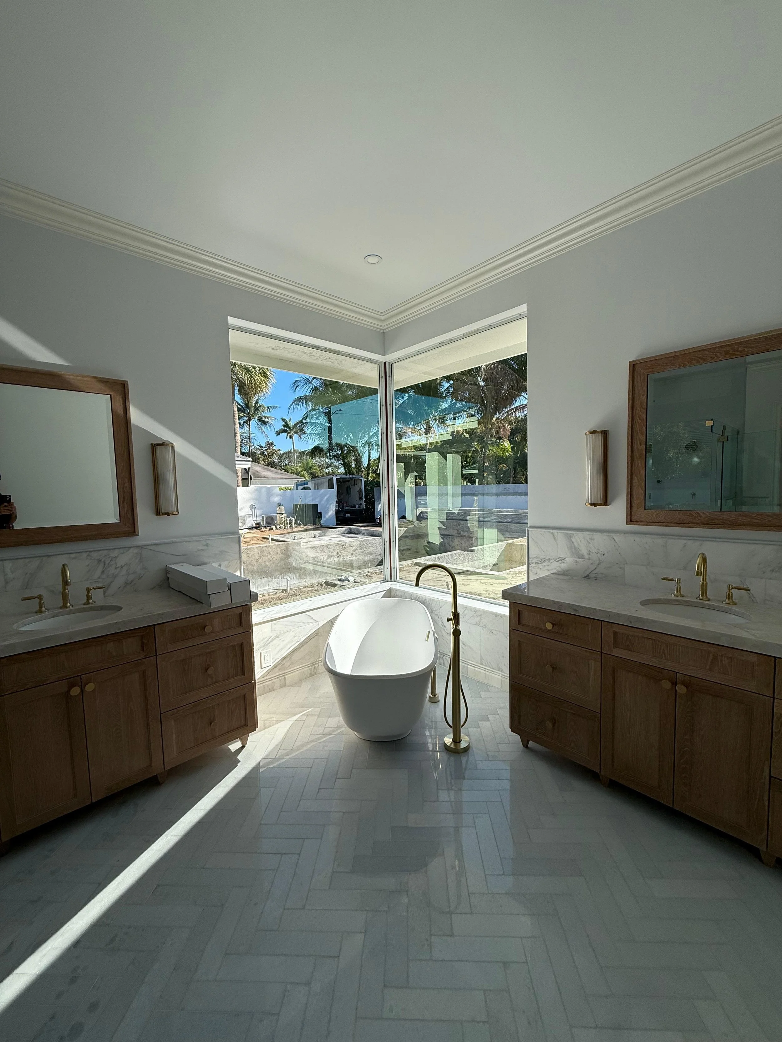 Modern bathroom with double vanity sinks, wooden cabinets, a freestanding bathtub, and large corner windows showing a view of palm trees.